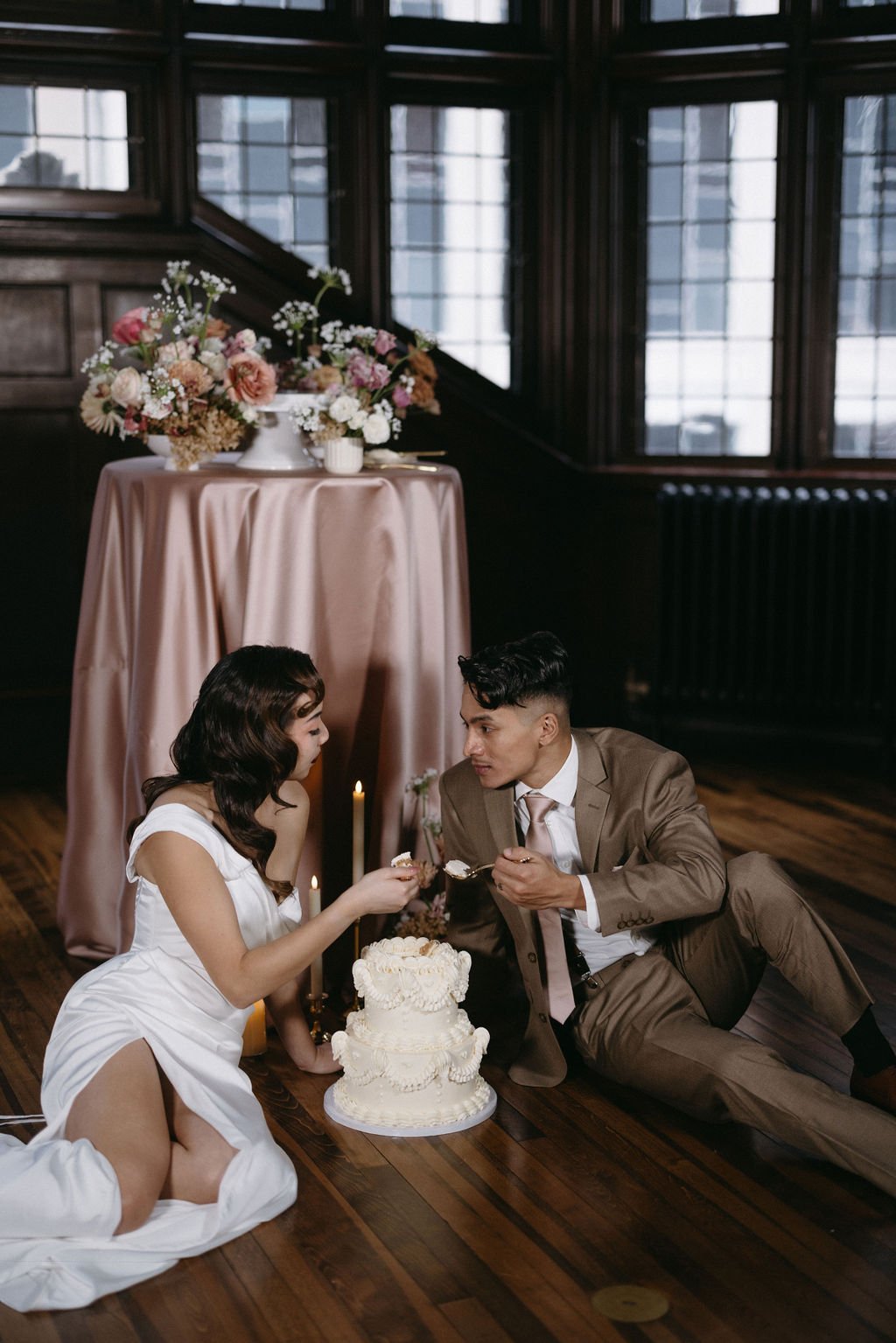 Bride and groom enjoying a casual cake-cutting moment sitting together on the floor beside a silk-draped table.   