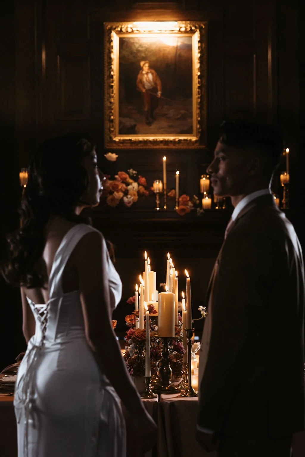 Moody wedding portrait of a couple lit by warm candlelight in front of an antique fireplace backdrop.