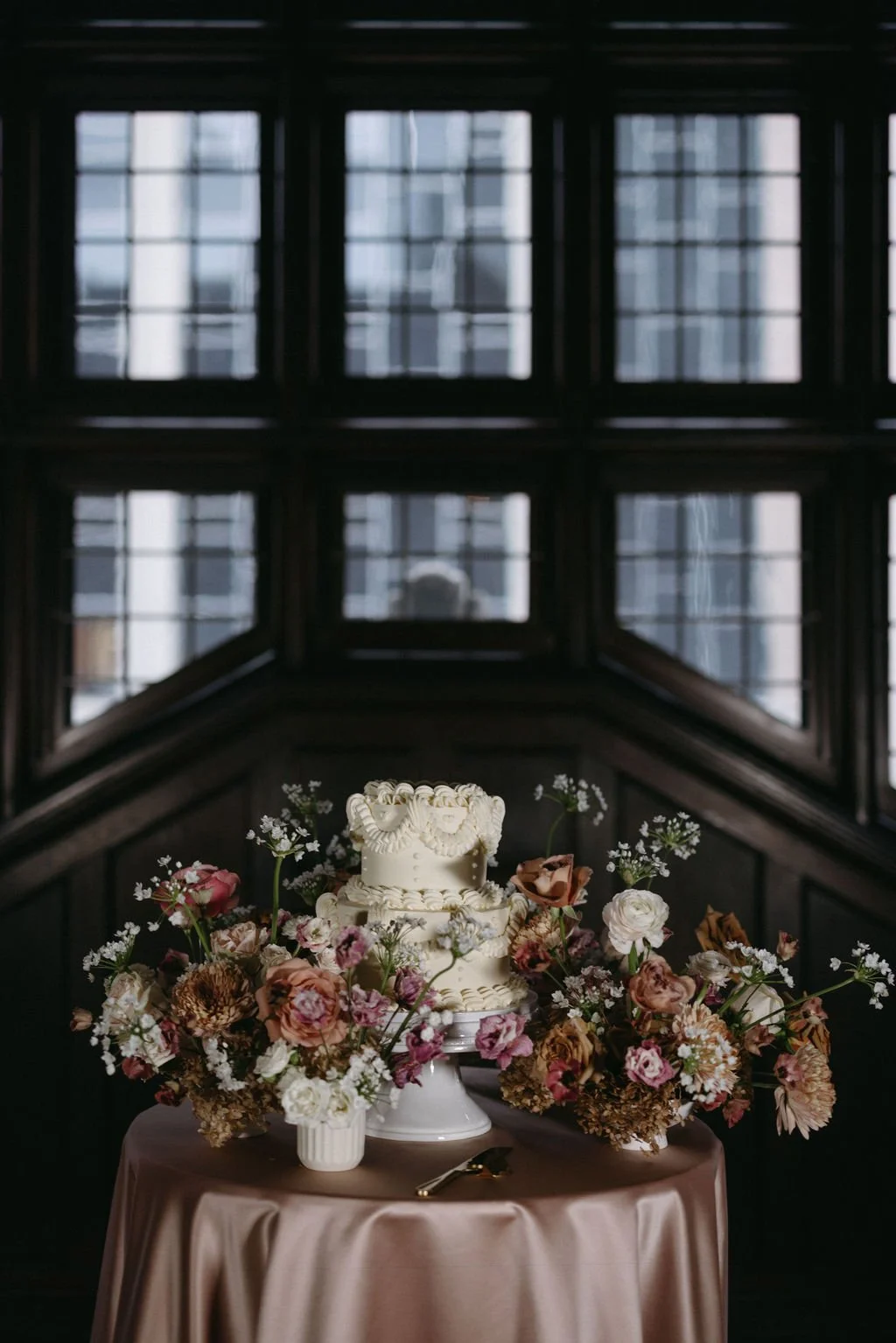 Elegant white textured wedding cake framed by rich mauve florals in front of an antique wood bay window.   