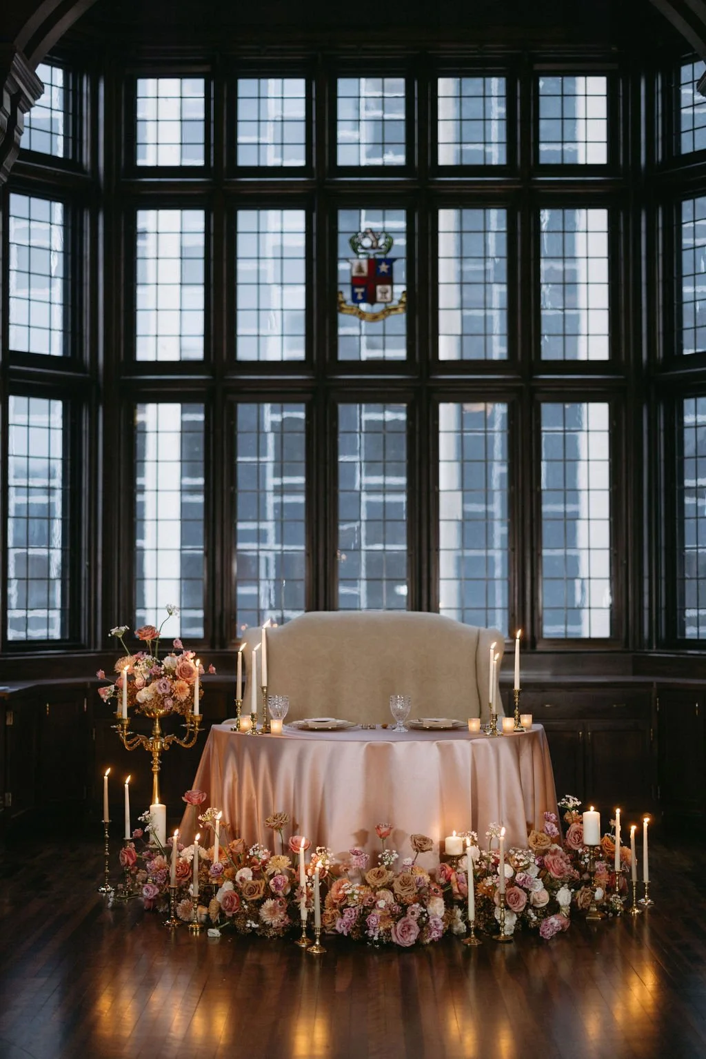 Intimate sweetheart table draped in blush linen, surrounded by floor candles beneath a dramatic leaded glass window.   