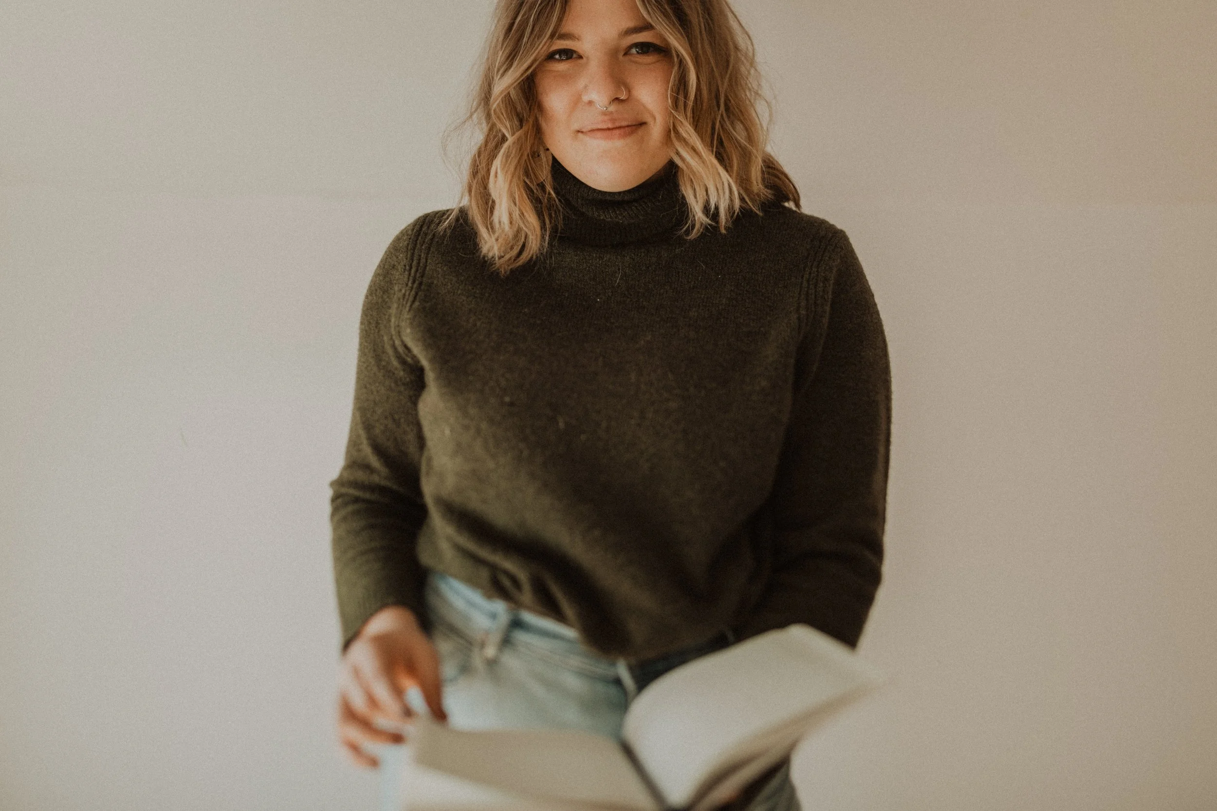A young woman with wavy blonde hair, wearing a black turtleneck sweater and light jeans, holding an open book in front of her. She is smiling slightly and standing against a plain light-colored wall.