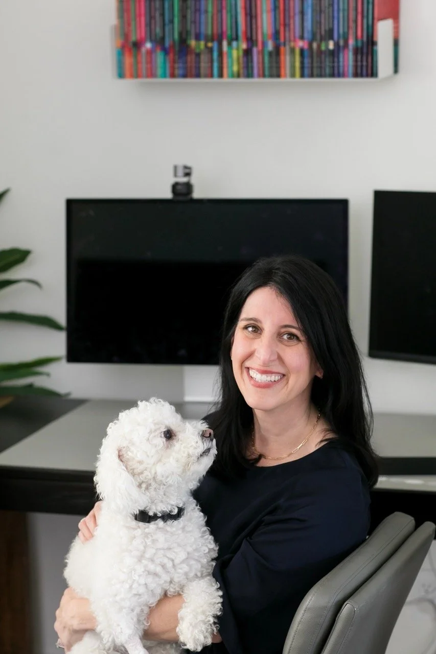 A photo of a smiling professional woman seated in front of her desk and computer monitors with a cute dog on her lap, looking approachable and competent