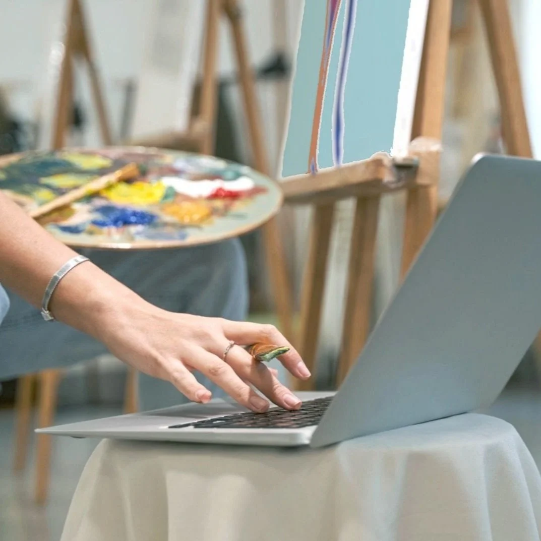 woman-seated-in-front-of-a-painting-easel, holding-a-paint-palette-in-her-left-hand and-typing-on-a-computer-with-her-right-hand-representing-a-creative-virtual-assistance-client