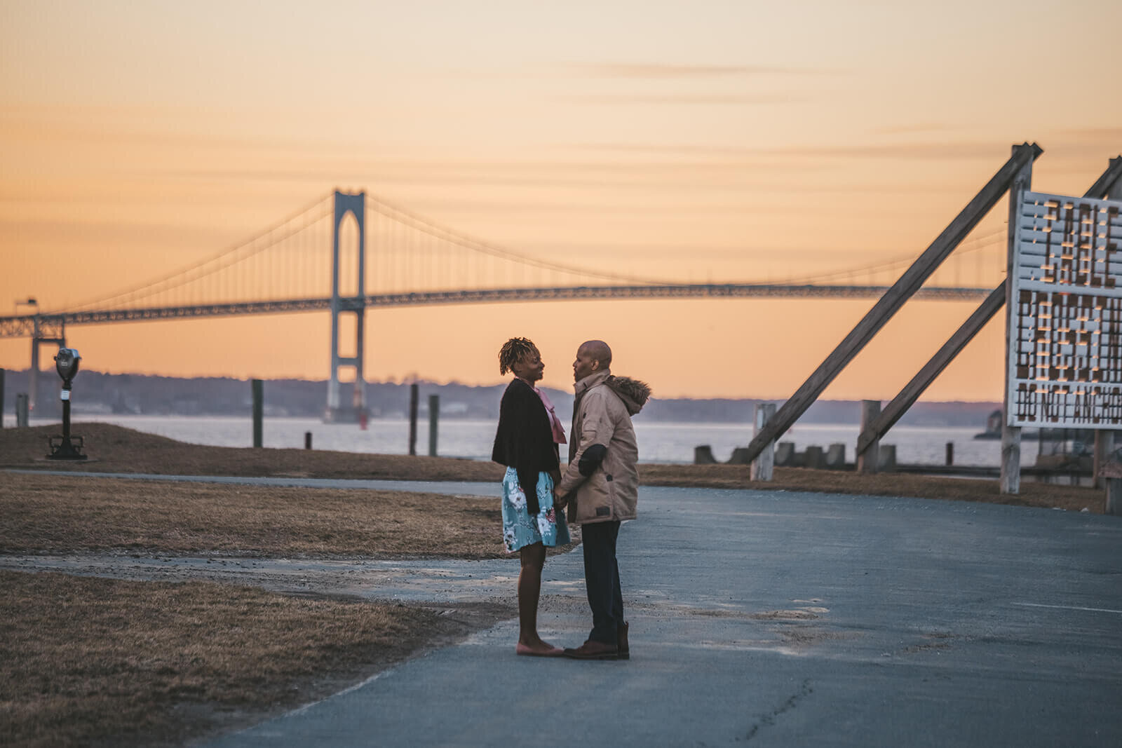  Couple stand in front of Newport Bridge at Fort Adams State Park at sunset 