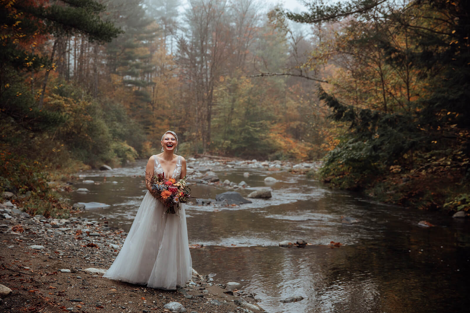  Bride laughs as she sees how beautiful the river is as she checks out the autumn colors surrounding her cabin in the woods in Vermont 