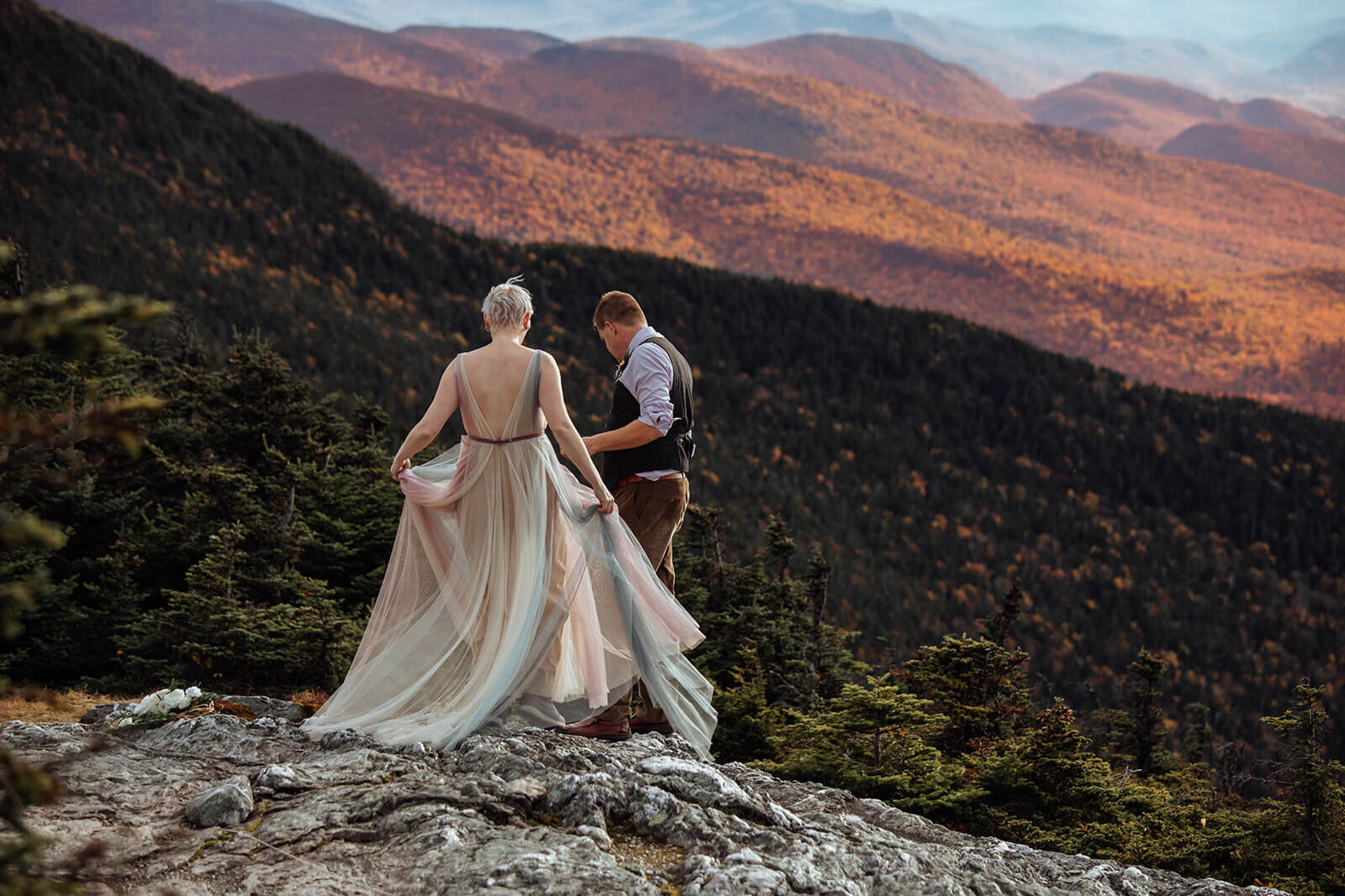  Brides dress flows in the wind  as she and the groom hike along summit of Mt. Mansfield near Stowe, Vermont after their elopement 