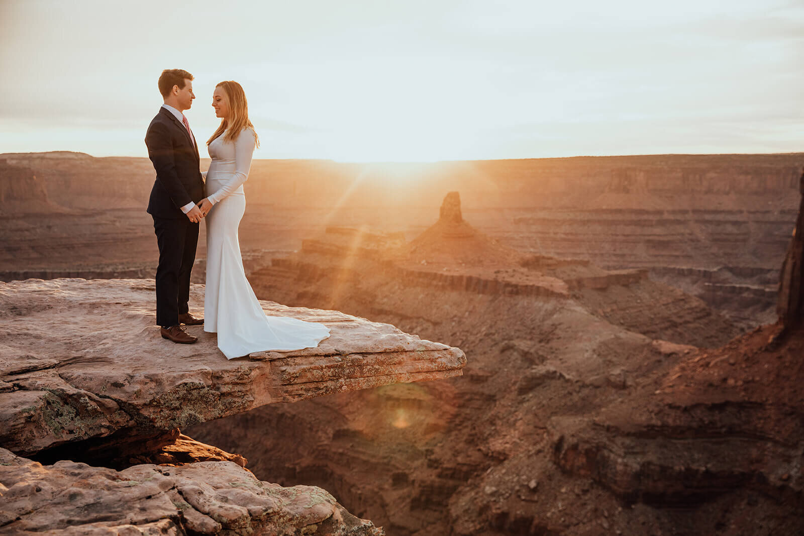  Couple celebrates anniversary and stands on cliff edge during sunset at Dead Horse Point State Park and the La Sal Mountains near Moab, Utah with incredible views and hiking. Utah elopement photographer 