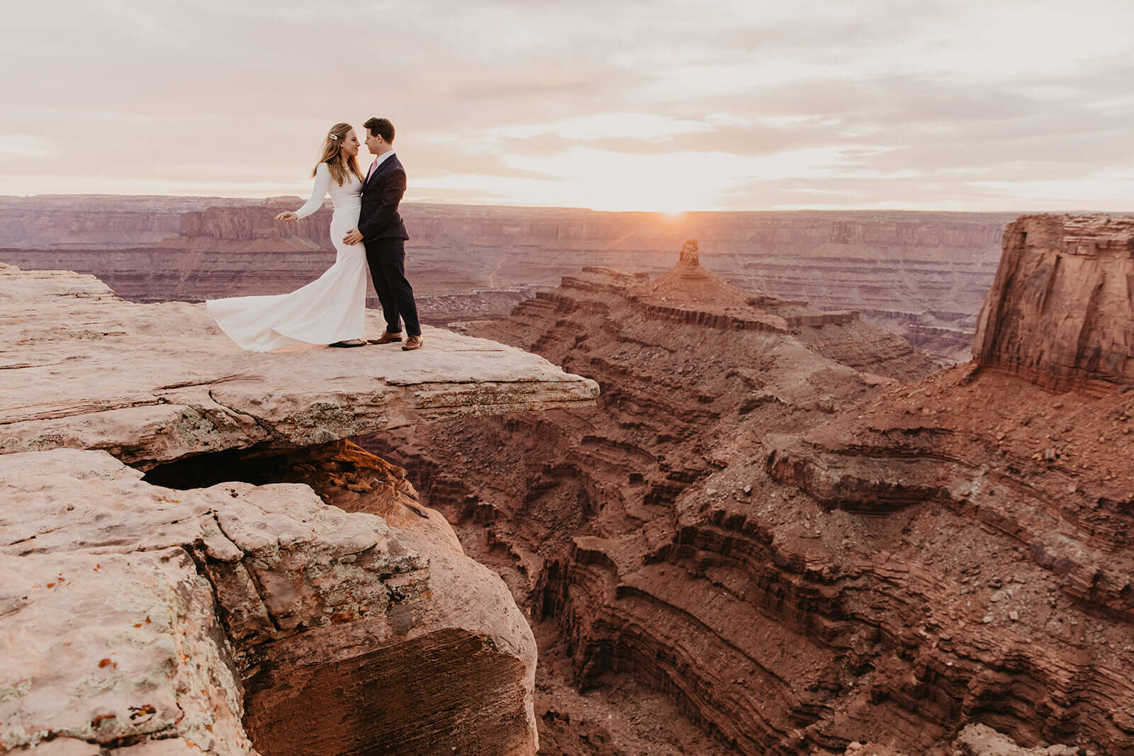  Couple standing on cliff edge during anniversary celebration at sunset at Dead Horse Point State Park and the La Sal Mountains near Moab, Utah with incredible views and hiking. Utah wedding photographer 
