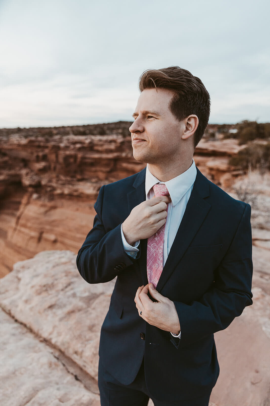  Groom straightens tie with standing on cliff edge during anniversary celebration at sunset at Dead Horse Point State Park and the La Sal Mountains near Moab, Utah with incredible views and hiking. Utah wedding photographer 