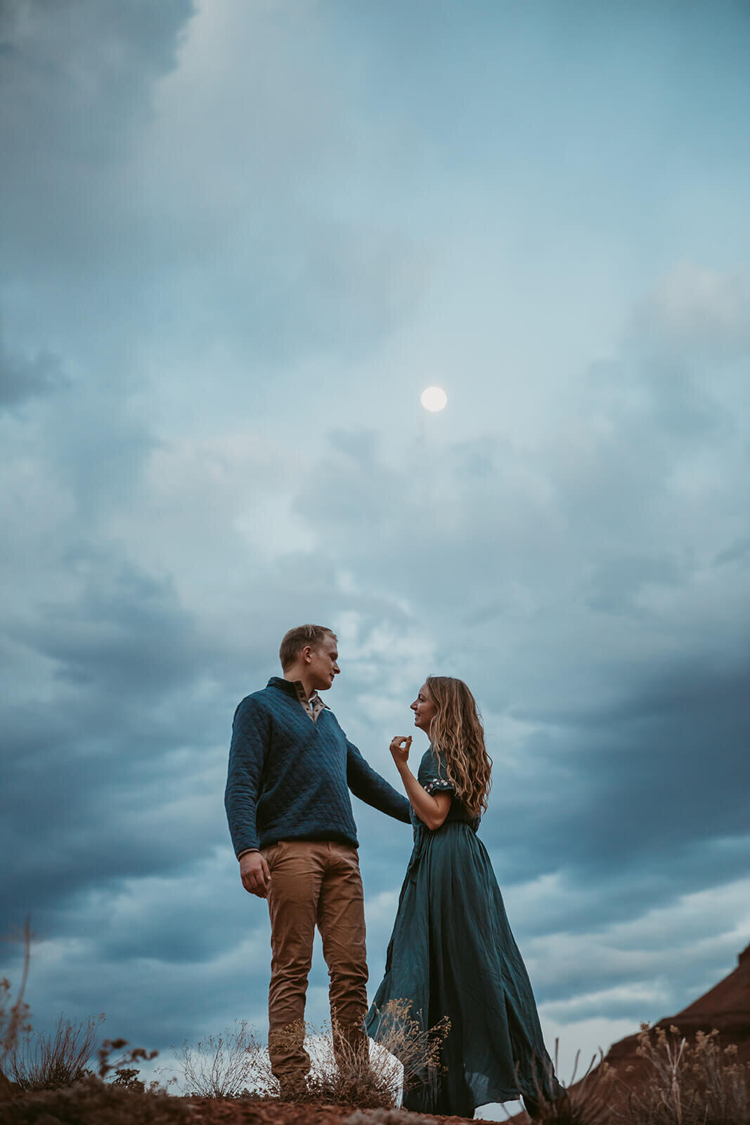  Couple are playful as a full moon rises over desert towers on public lands outside of Moab, Utah. Utah engagement photographer 