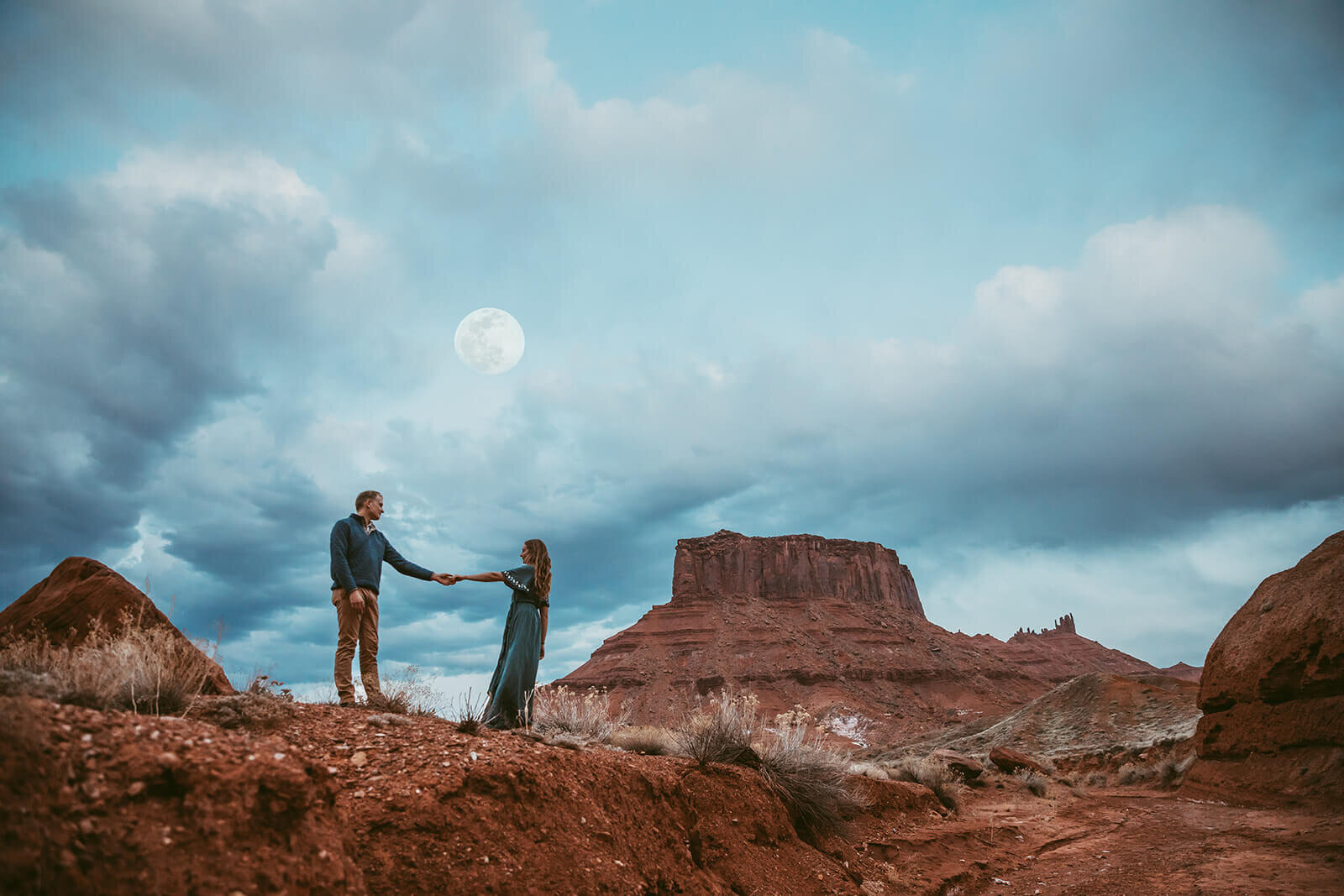  Couple are playful as a full moon rises over desert towers on public lands outside of Moab, Utah. Utah engagement photographer 