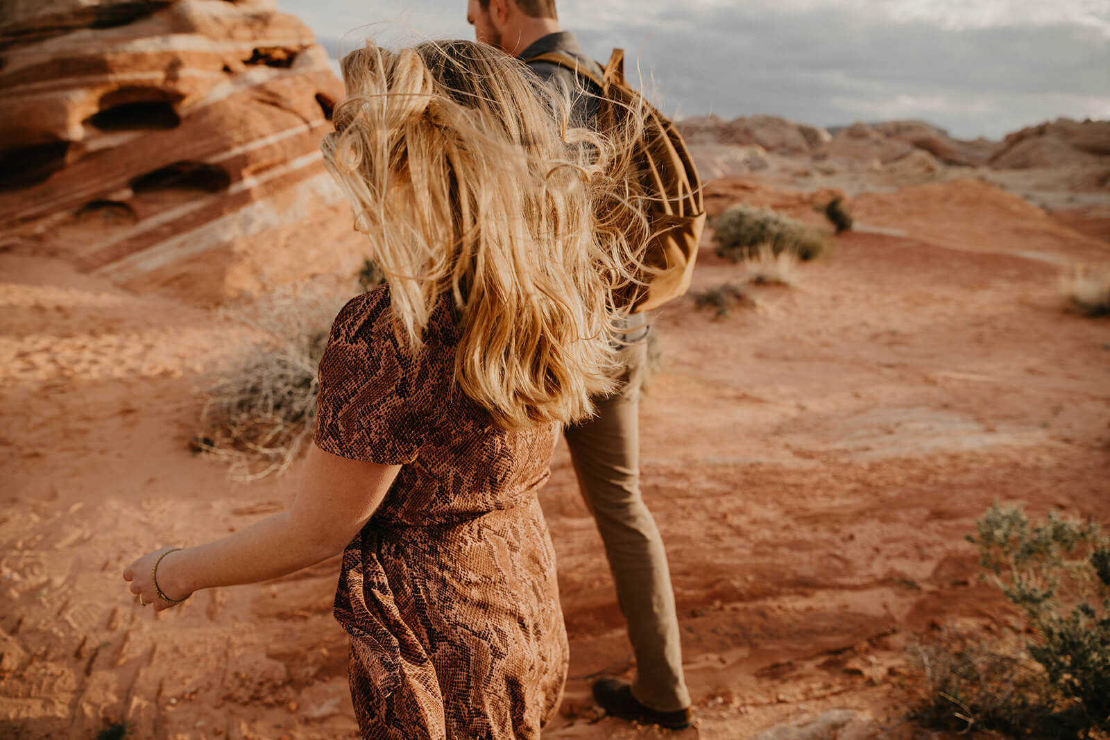  Hair blows in the wind as couple hikes through canyon in Valley of Fire State Park next to Lake Mead, outside of Las Vegas, Nevada. Las Vegas engagement photographer 