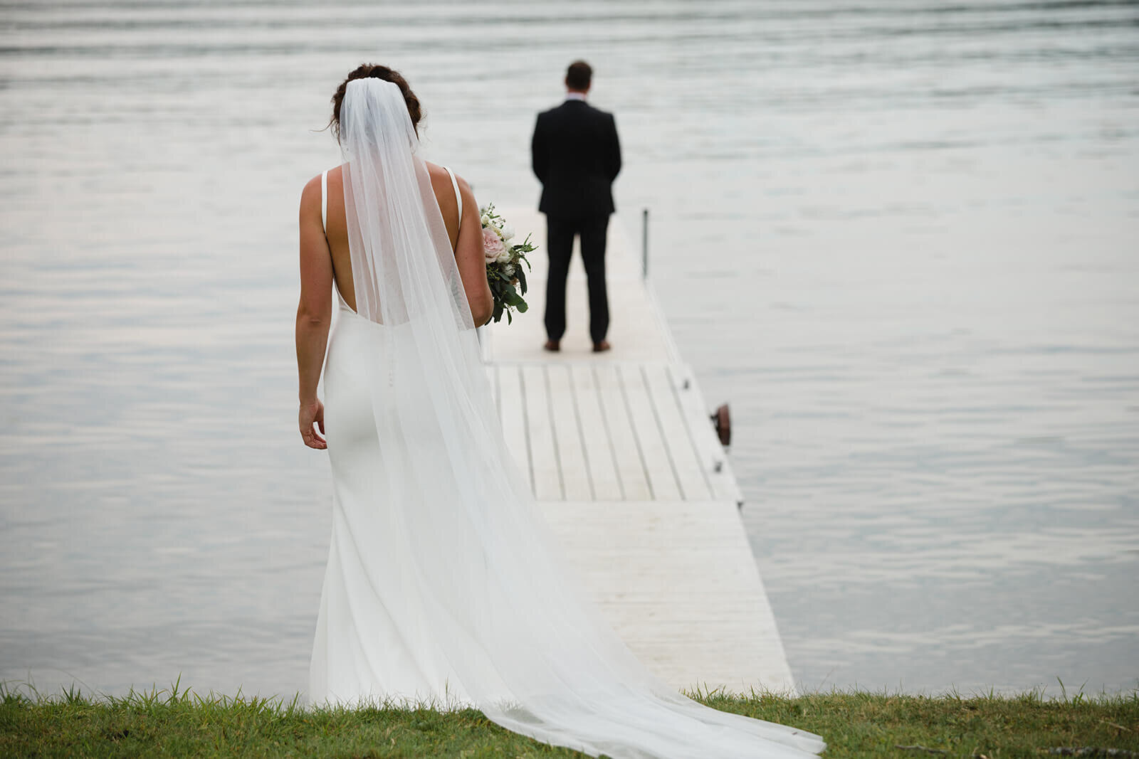  Bride walks toward groom for first look on dock. Small outdoor wedding on a lake in Maine. 
