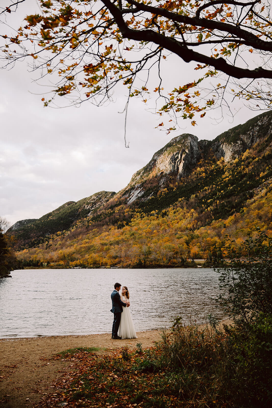  Couple explores a lake in the rainy misty weather in Franconia Notch State Park in New Hampshire after their elopement ceremony. New Hampshire elopement packages. 