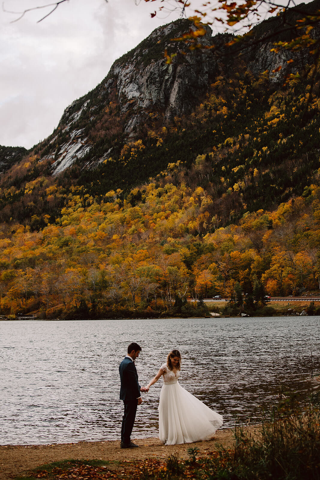  Couple explores a lake in the rainy misty weather in Franconia Notch State Park in New Hampshire after their elopement ceremony. New Hampshire elopement packages. 