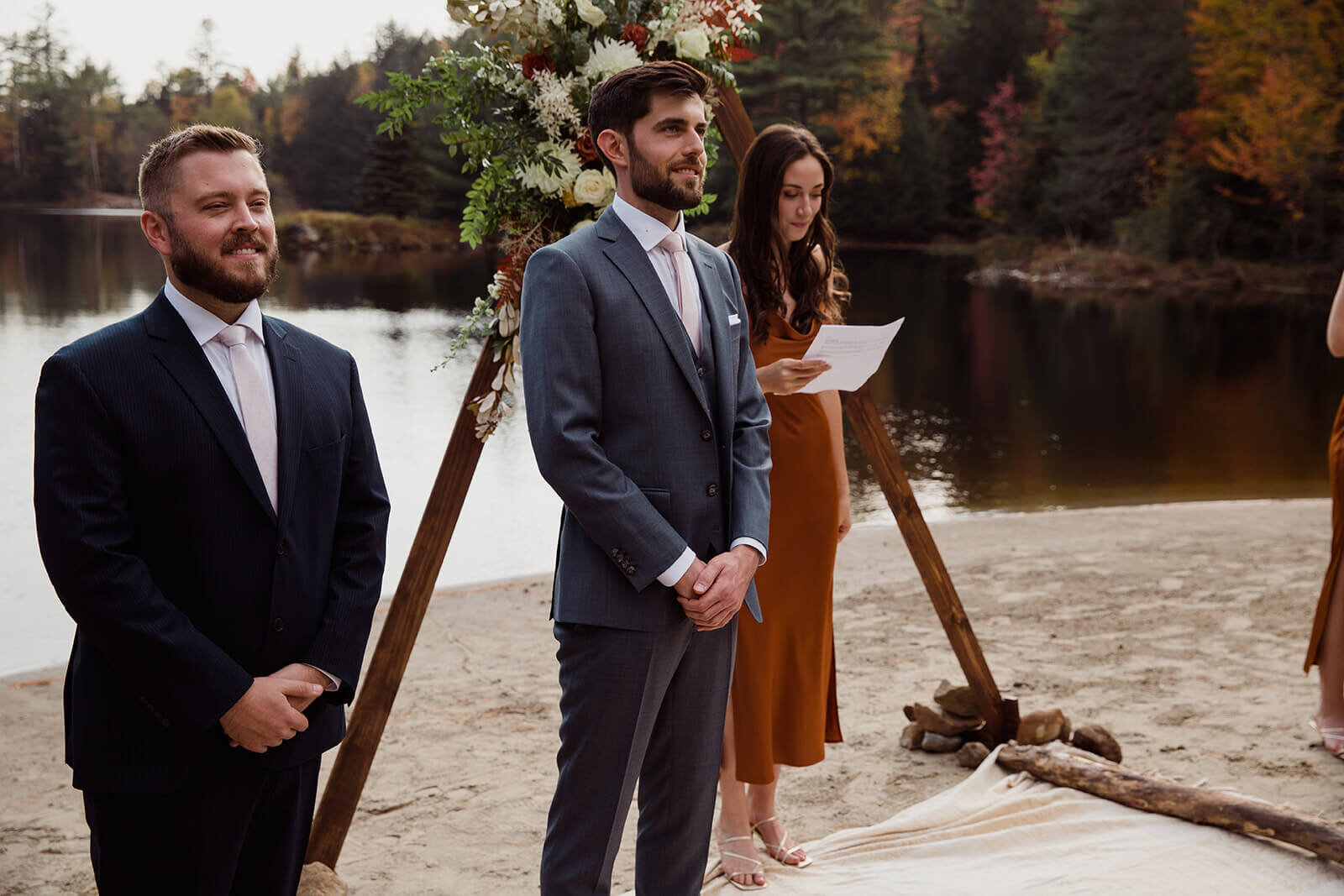  Groom sees bride walking down the aisle on the lake beach during their fall wedding in the Adirondacks in Upstate New York. 