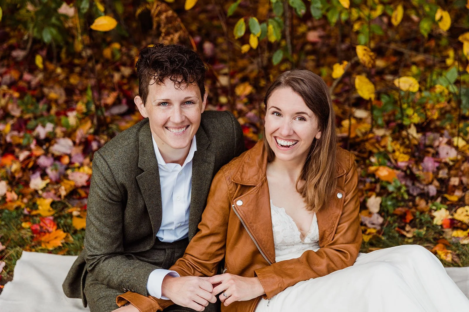 Two brides holding hands while sitting on a blanket in an autumn park with fallen leaves, while eloping in Vermont.
