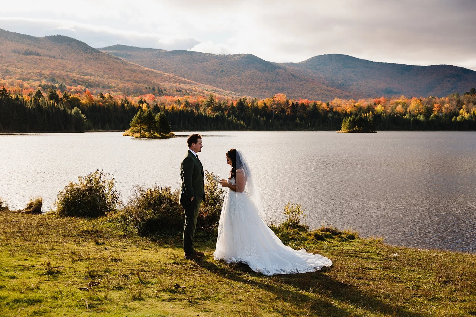 A bride and groom standing on the grass near a lake during their wedding, with Vermont's Green Mountains and colorful autumn trees in the background.