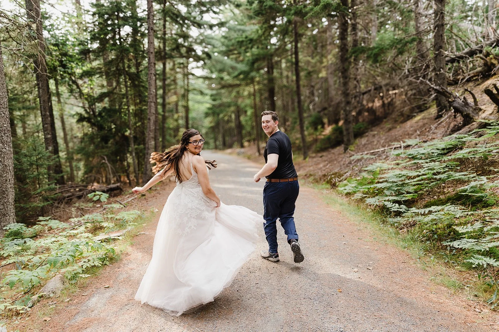 A joyful couple, dressed in wedding attire in Acadia National Park in Maine, running along a forest trail surrounded by tall trees and green foliage.
