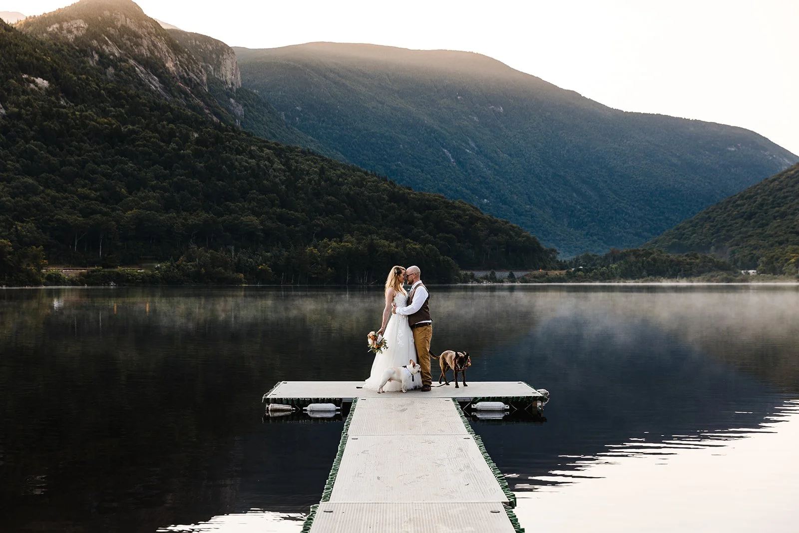 A bride and groom standing on a dock by a lake, holding hands and touching foreheads, with two dogs nearby. The White Mountains in NH in the background.