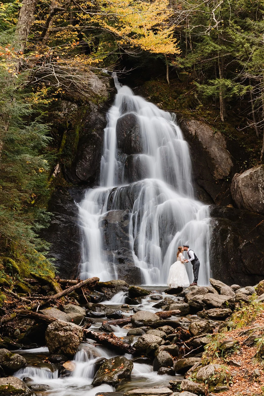 A bride and groom in wedding attire in Vermont sharing a kiss in front of a cascading waterfall in a lush forest.
