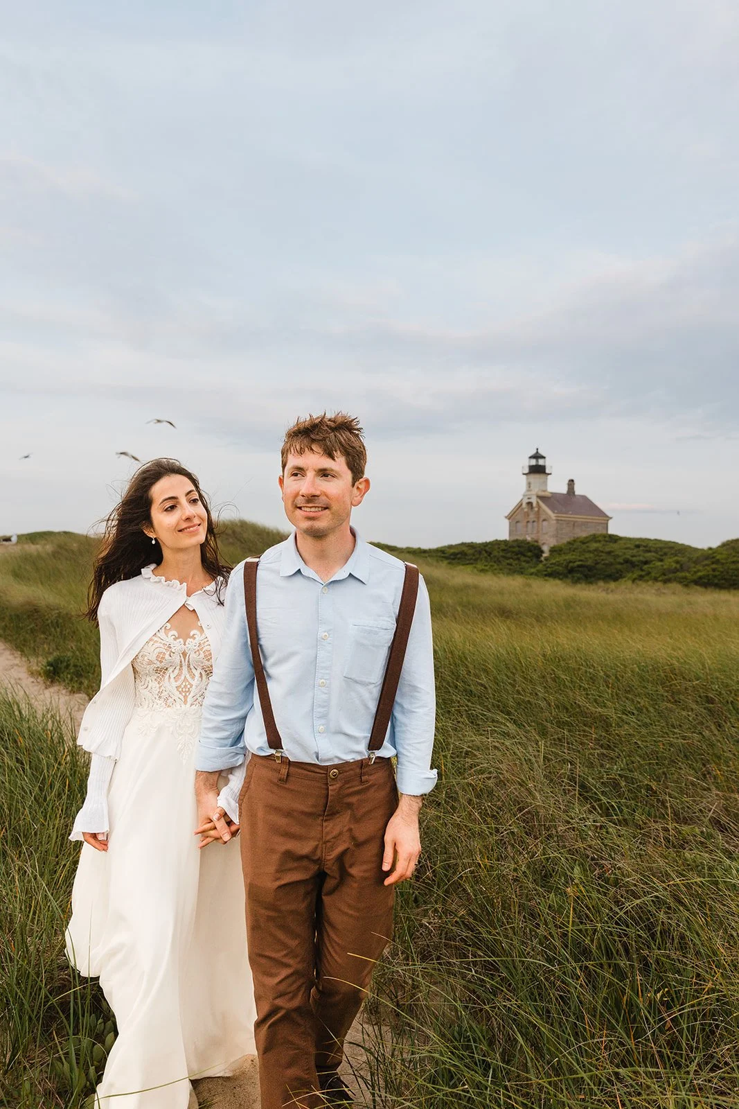 A couple walking hand in hand through grassy dunes with a lighthouse in the background during daytime while eloping on Block Island.