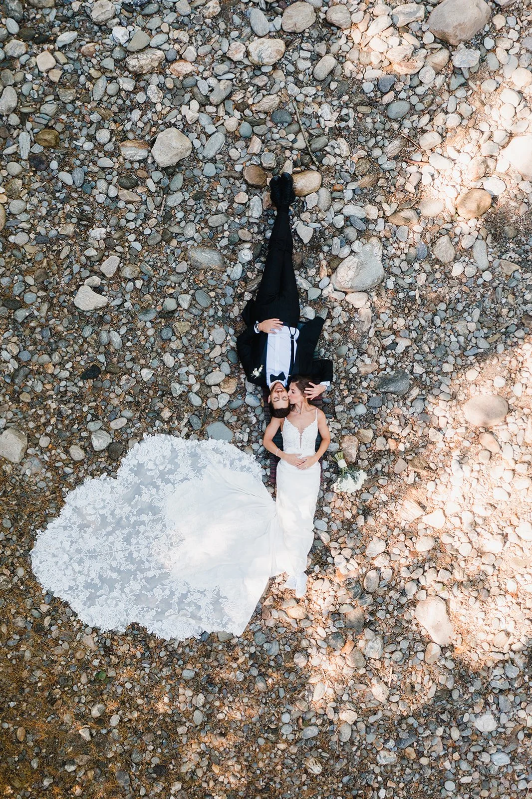 A bride in a white wedding gown and a groom in a black tuxedo lying on rocky ground in Vermont, seen from above, with the bride wearing a lace gown and holding a bouquet of white flowers.