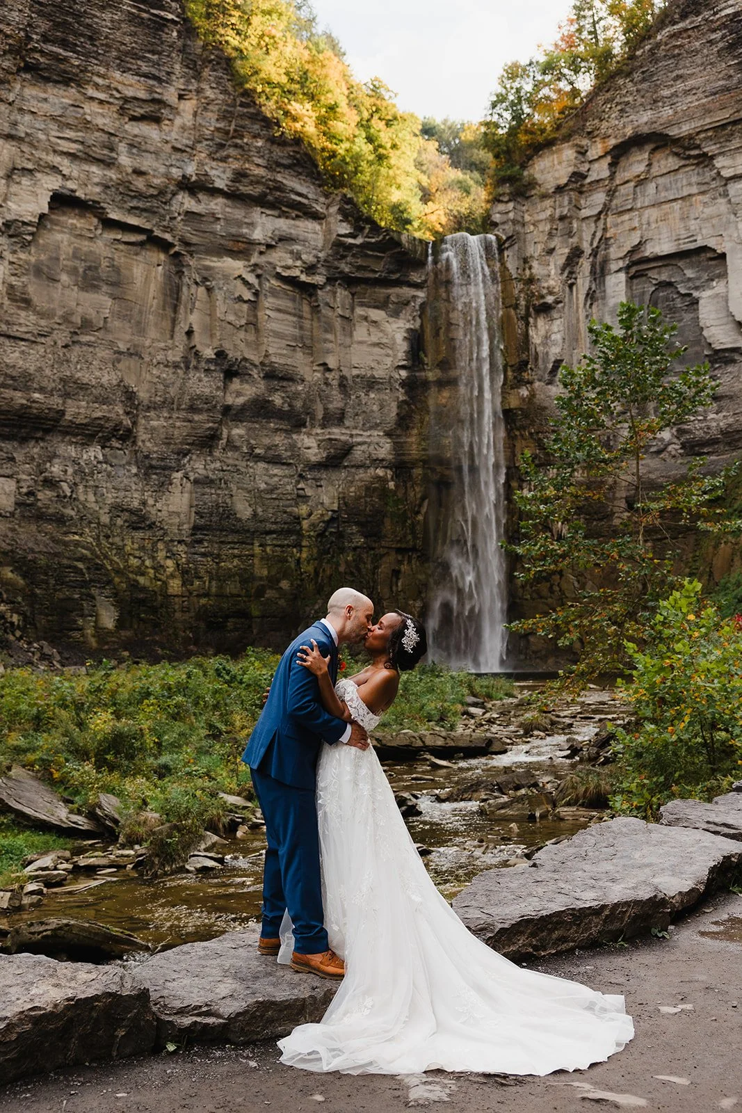 A bride and groom kissing in front of a waterfall in the Finger Lakes of Upstate NY in a natural outdoor setting, with greenery and rocks surrounding them.