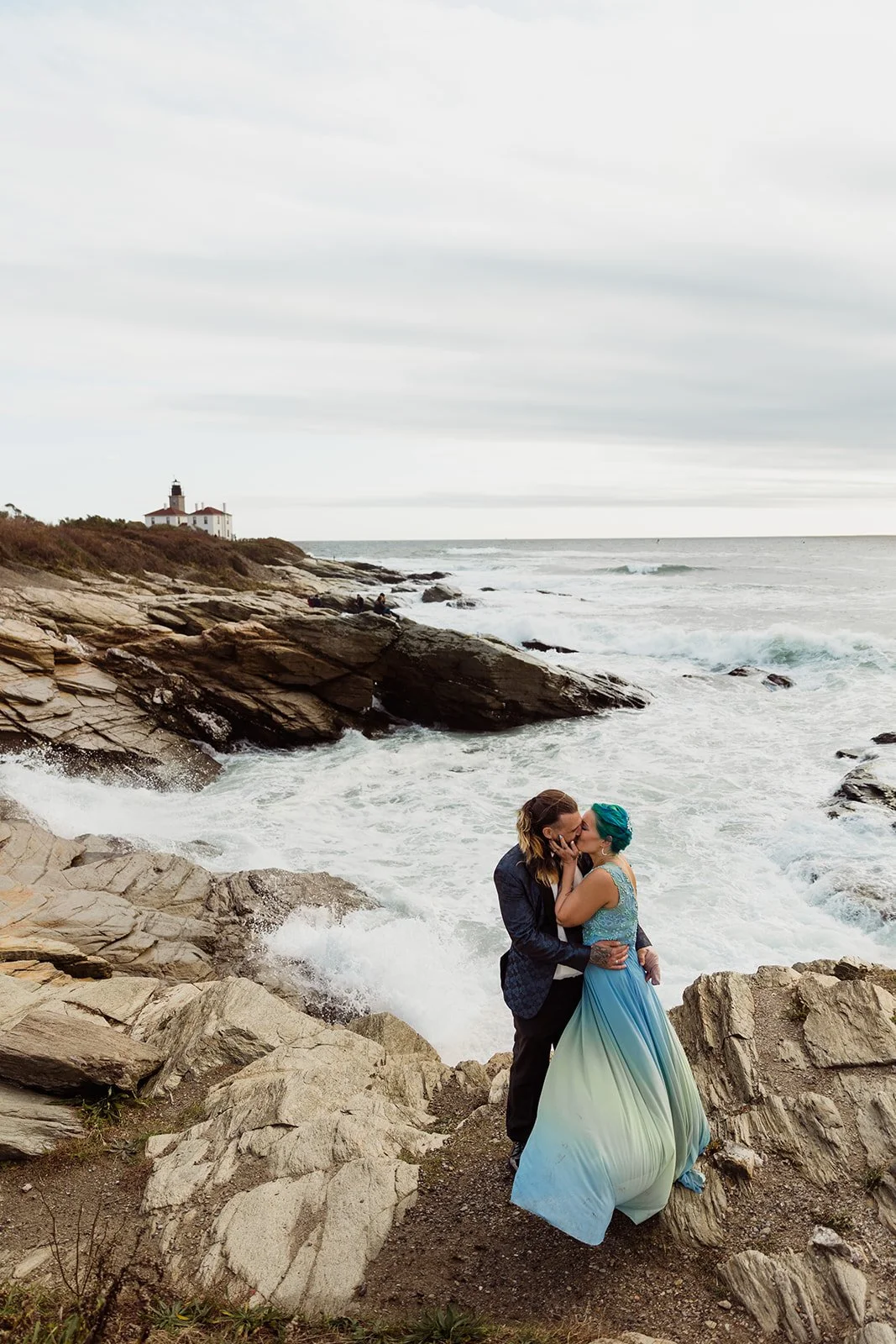 A couple kissing on rocky coast in RI while eloping near the ocean with lighthouse in the background, one dressed in a suit and the other in a blue wedding dress with teal hair.