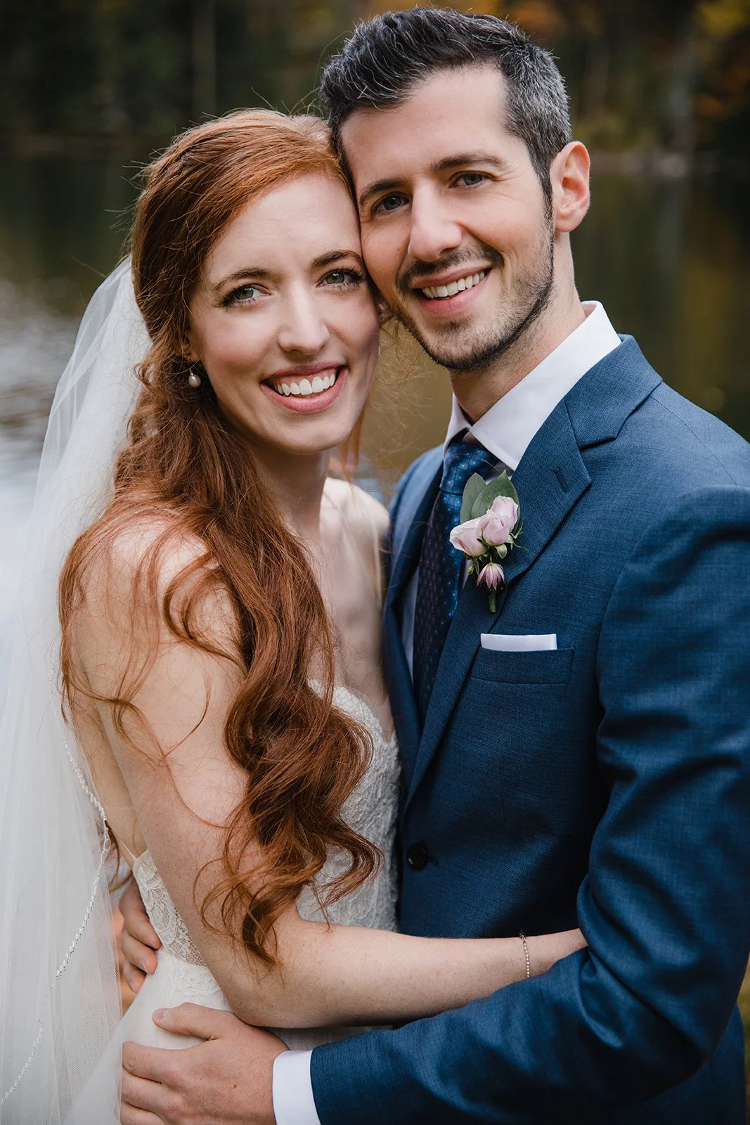 A happy bride and groom smiling and embracing outdoors near a body of water, with trees showing fall colors in the background in NH.