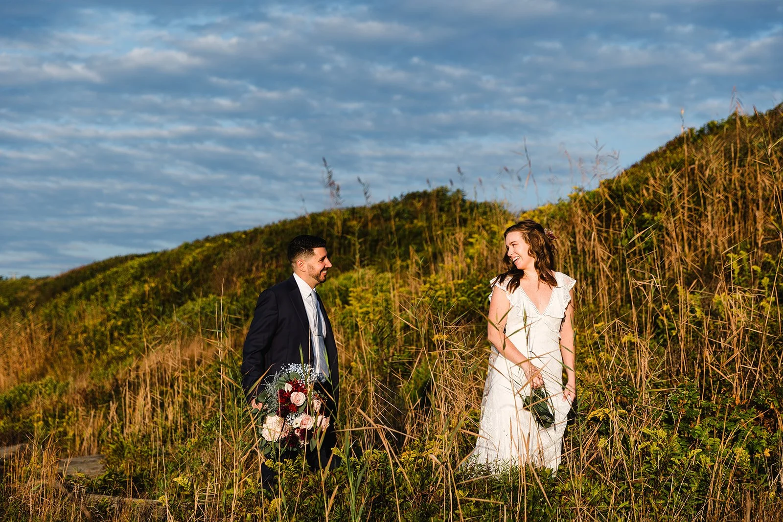 A couple dressed in wedding attire standing in tall grass on a hillside, on the coastal cliffs of Rhode Island, smiling at each other, with a cloudy sky above.