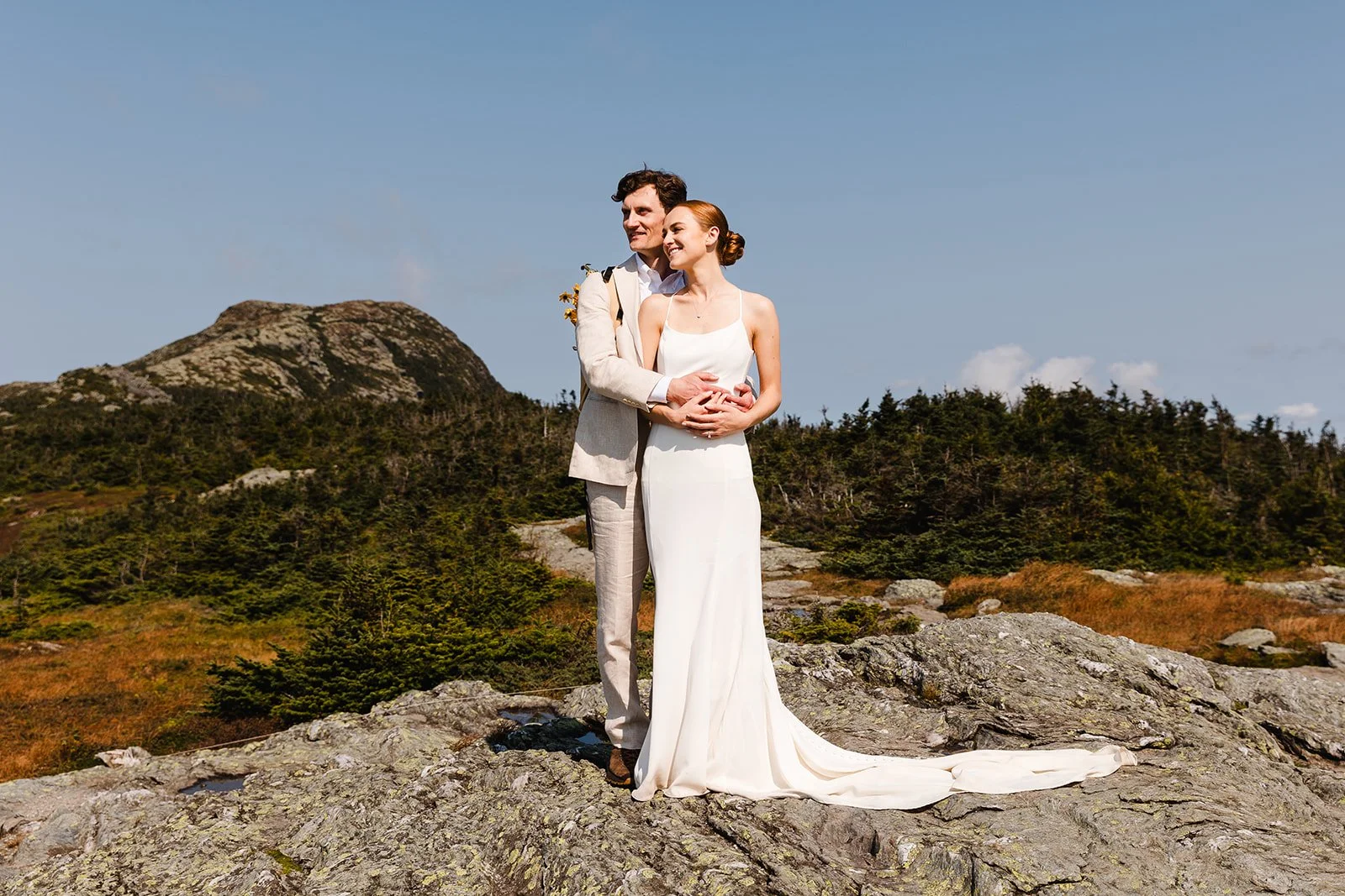 A bride and groom standing on a rocky landscape on the tallest mountain in VT with mountains and green trees in the background. The bride is wearing a white wedding dress and the groom is in a light-colored suit.