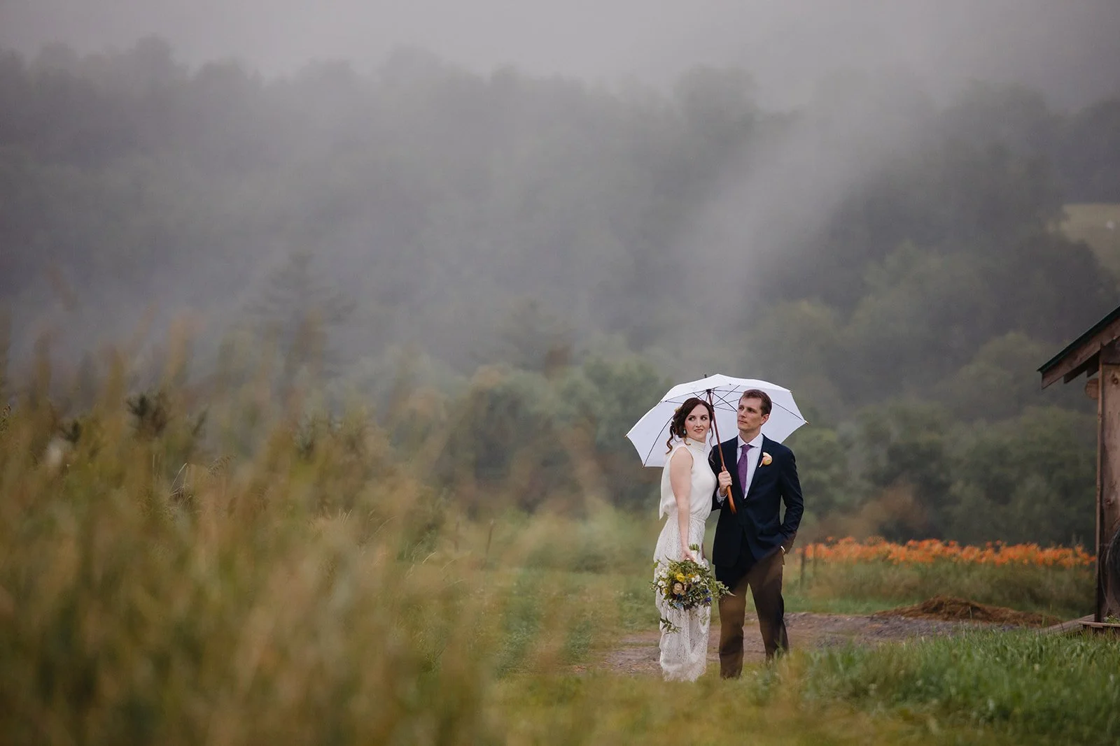 A bride and groom standing in a grassy field on a rainy day, sharing an umbrella and looking into the distance, with trees and a wooden structure in the background in VT.