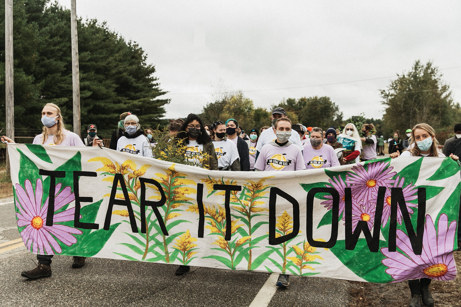 image of a crowd of people in t shirts that say "no coal no gas" walking behind a big banner that says "Tear it down." The people are carrying plants and buckets of soil