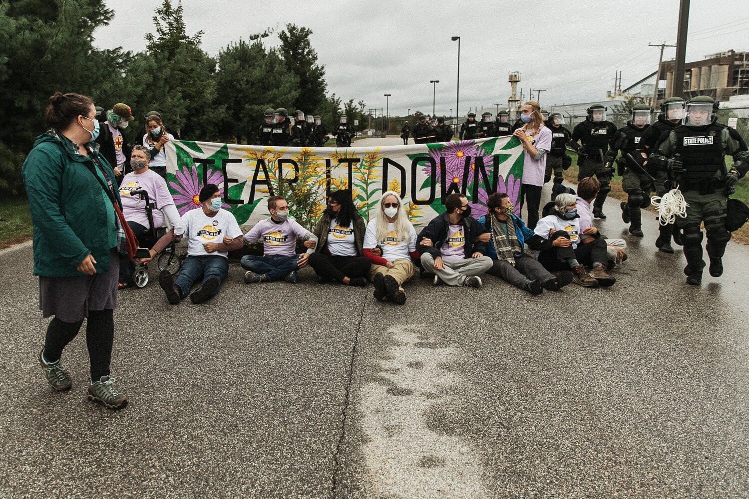 image of a group of individuals sitting on concrete in front of a banner that has flowers on it and the text "Tear it down." The coal plant in Bow, NH is in the distance and cops in riot gear are approaching the group.