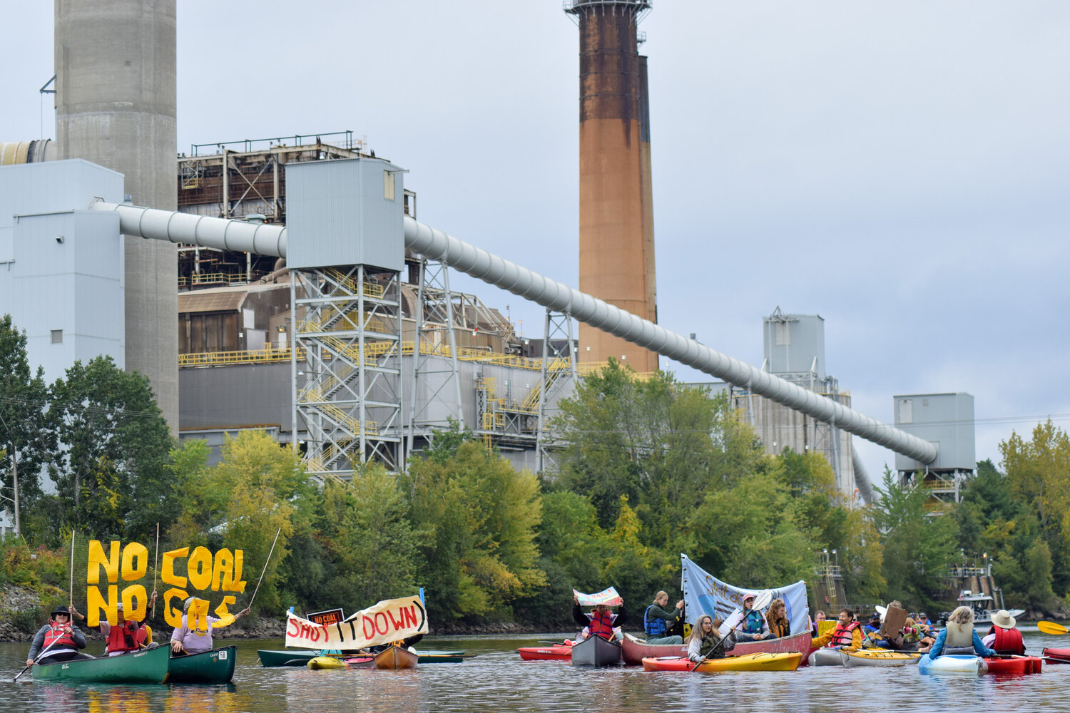 image of a group of kayaks and canoes in the Merrimack River, holding various signs that say "shut it down" and "no coal no gas." The big looming coal plant is in the background