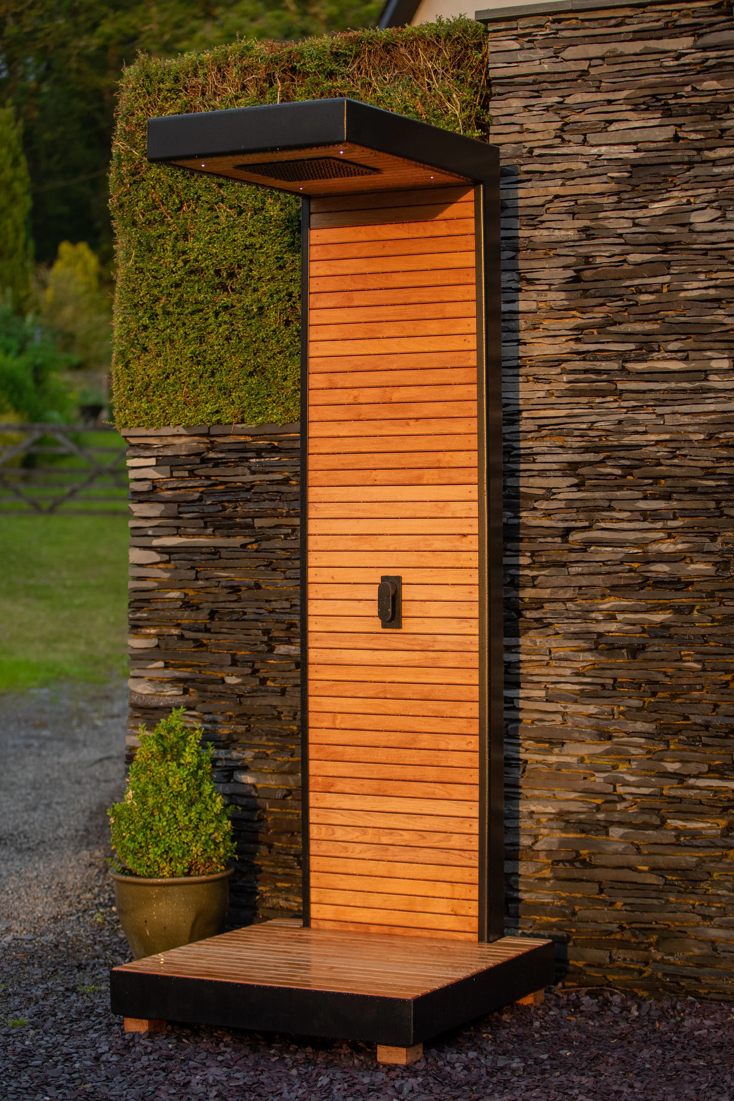 An outdoor wooden shower with a black metal frame against to a stone wall, with a small potted shrub nearby, in a garden setting during sunset.
