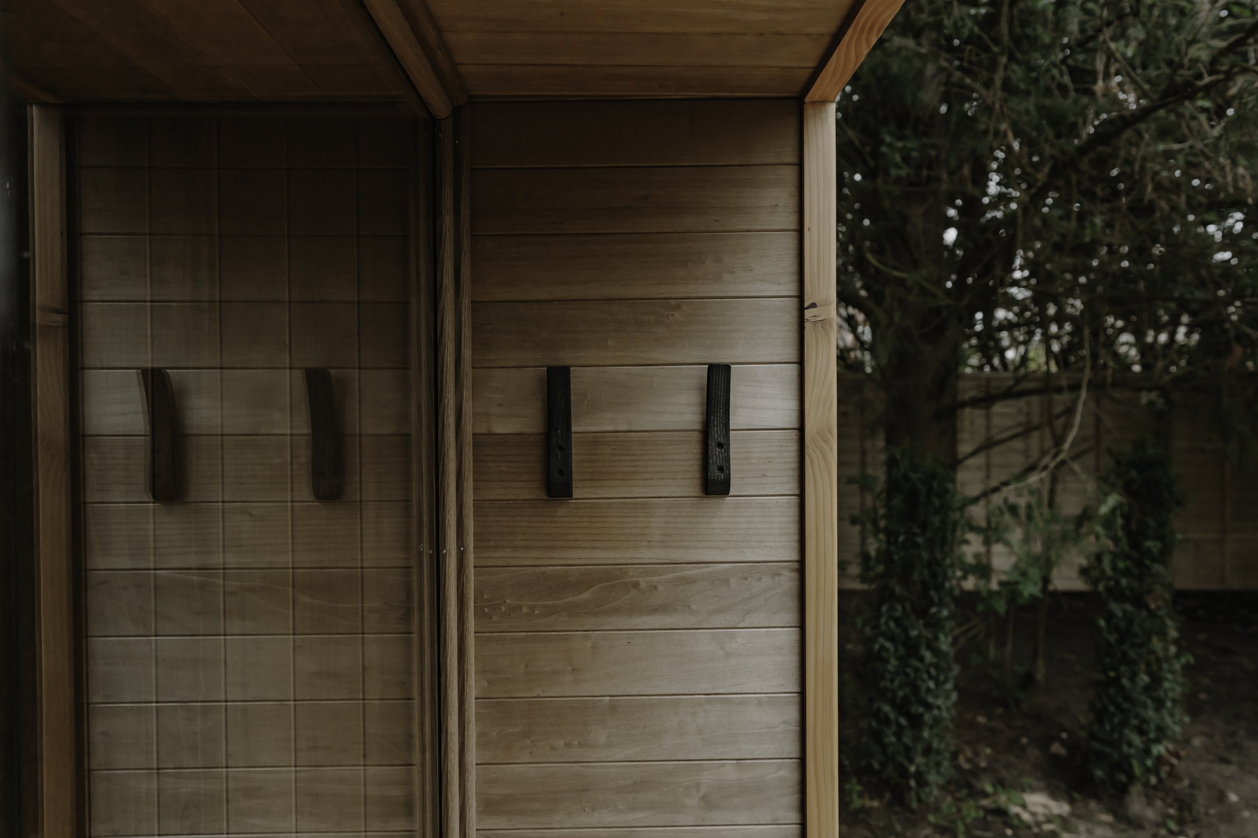 Interior of a wooden outdoor sauna porch with black wooden clothes hooks on the wall and a glass door, outside trees and greenery visible.