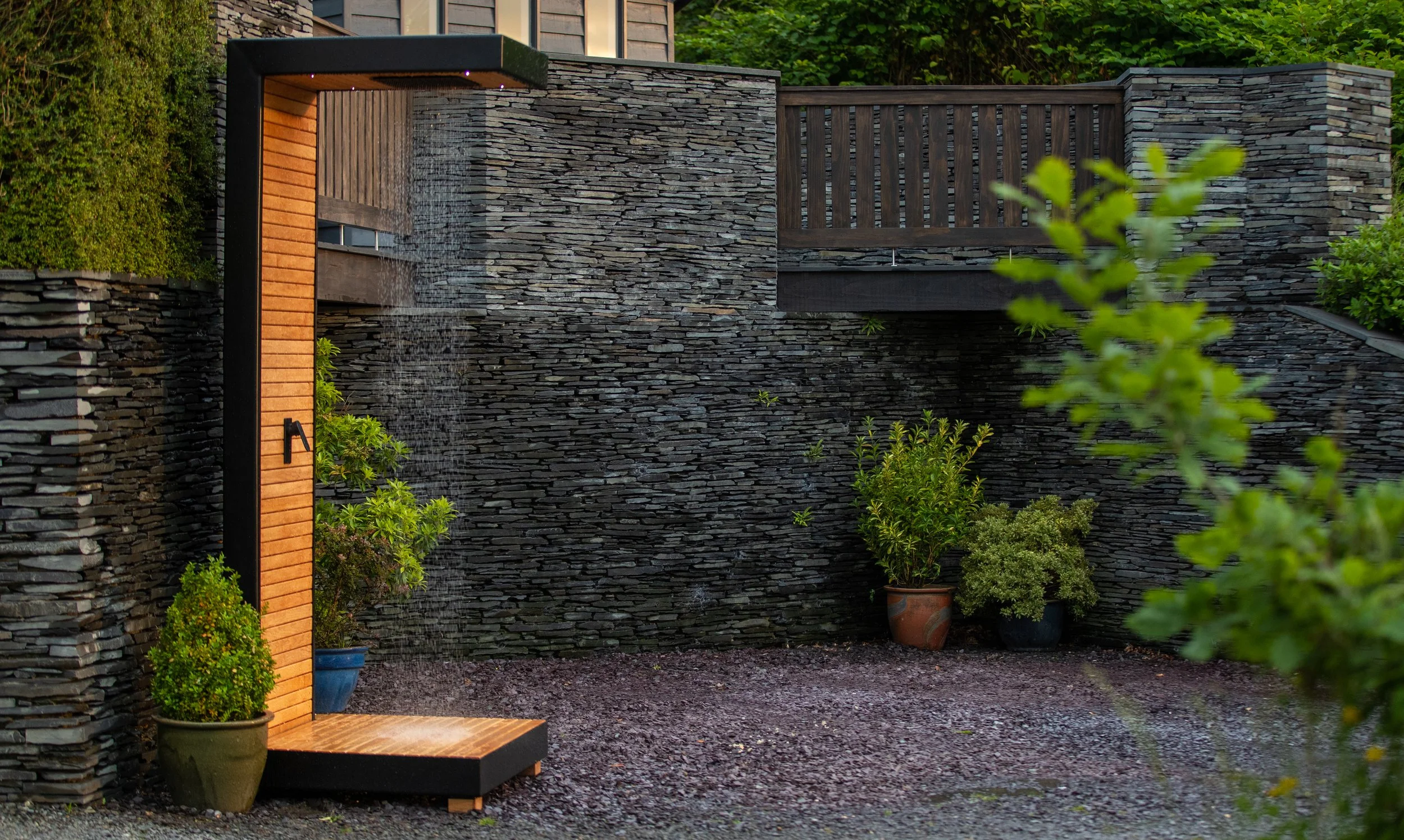 Exterior patio shower area with a modern black and natural wood-cladded outdoor shower featuring a rain shower head, set against a dark slate stone wall with potted plants nearby.