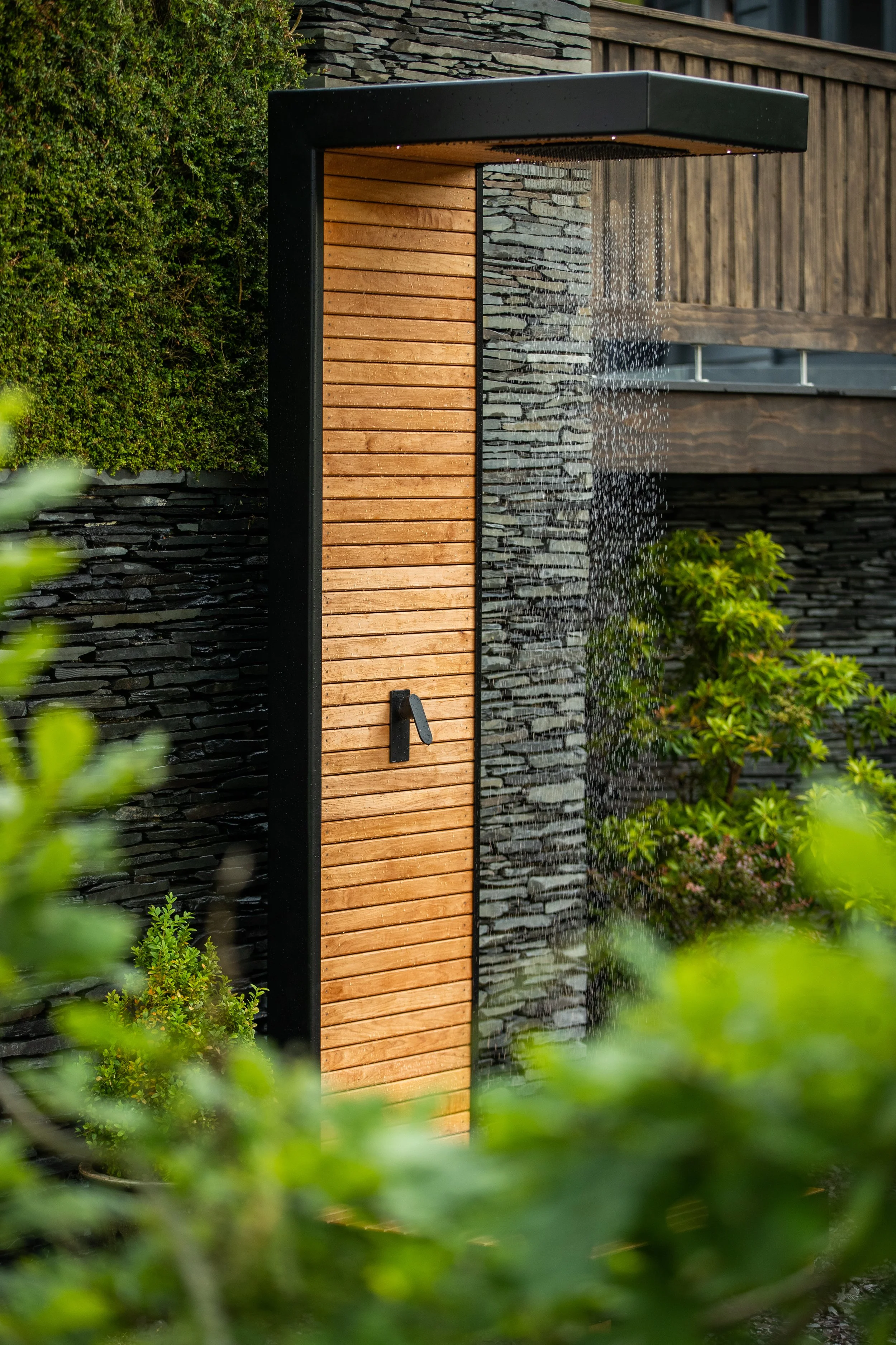 Outdoor shower with wooden panelling and black metal frame seen through two green hedges with water cascading down from a rainforest shower head with external slate wall of a house behind. 