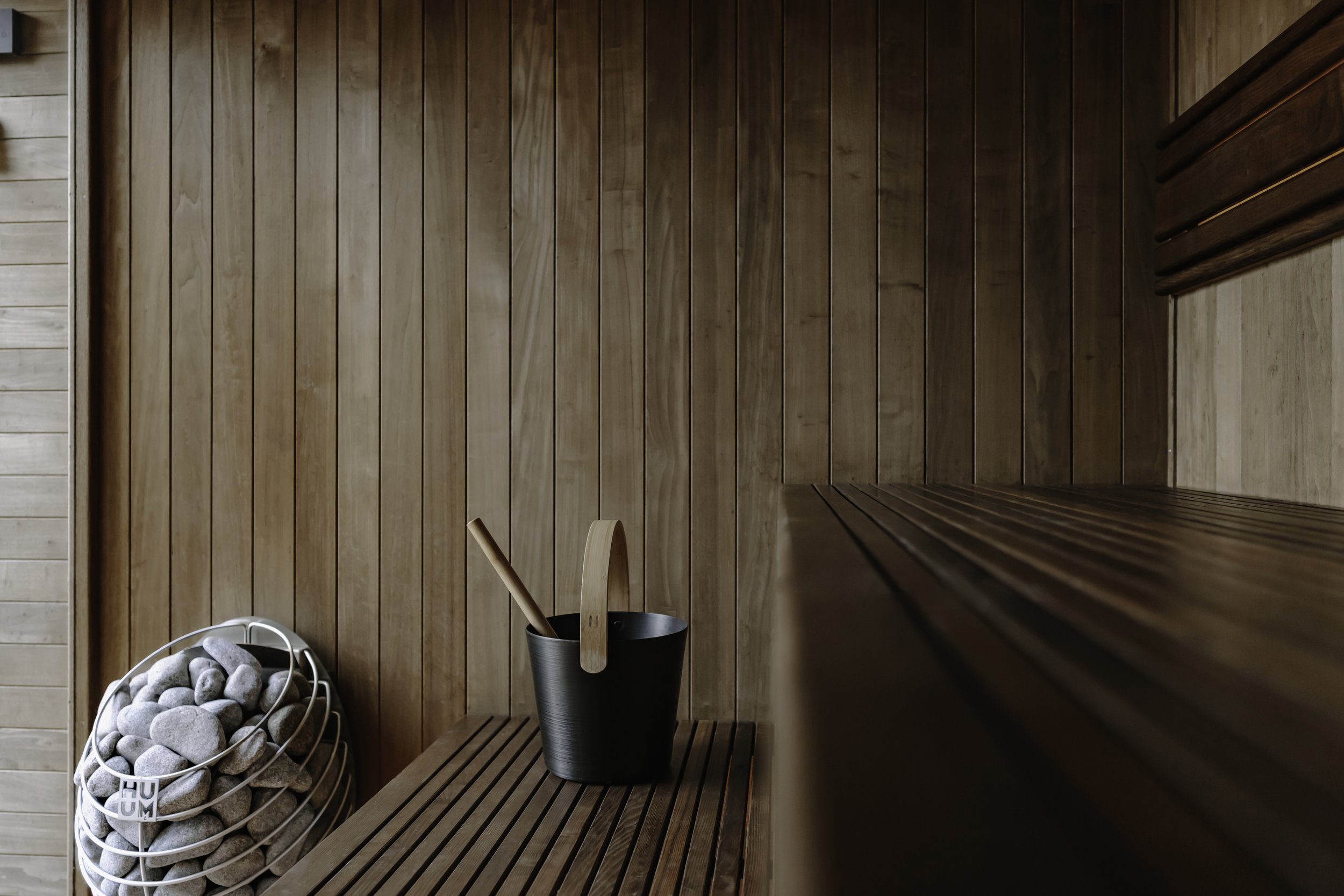 Interior of a wooden sauna with two teirs of benches, a black bucket with a wooden ladle, and an electric sauna stove with stones labeled "HUUM".