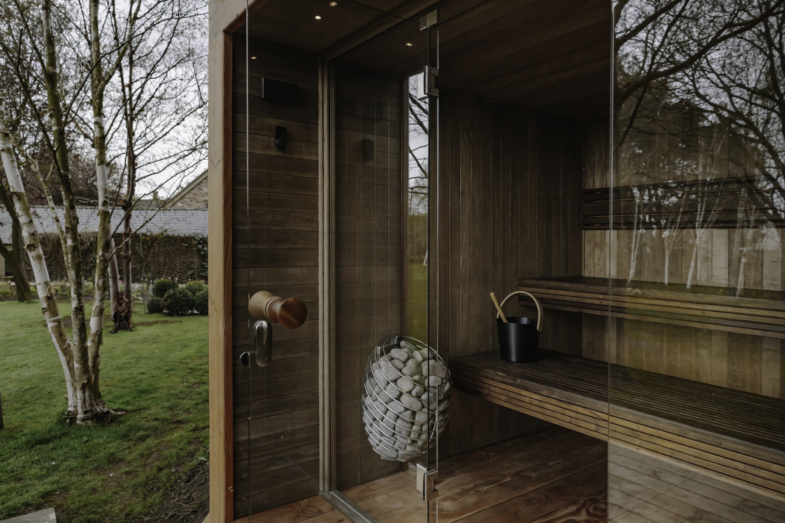 Interior of a wooden sauna with glass door, wooden benches, a bucket with a ladle, and an electric sauna stove, with an outdoor garden visible through the glass.