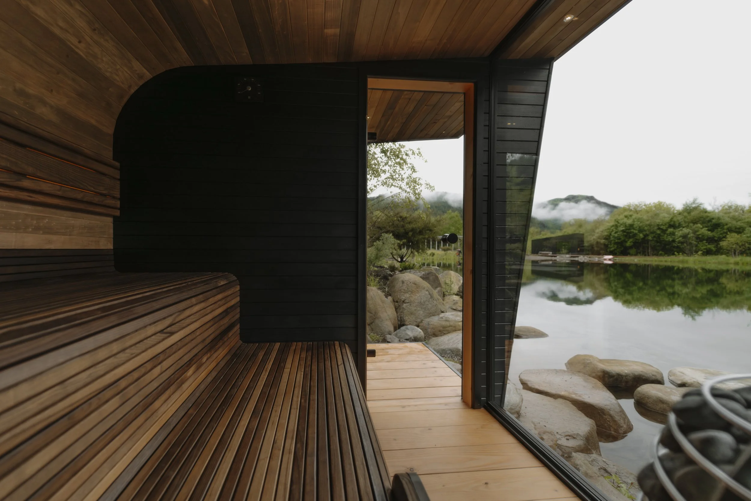 Interior of a black and naturally wooden-cladded sauna with a large glass landscape window overlooking a lake and green landscape.