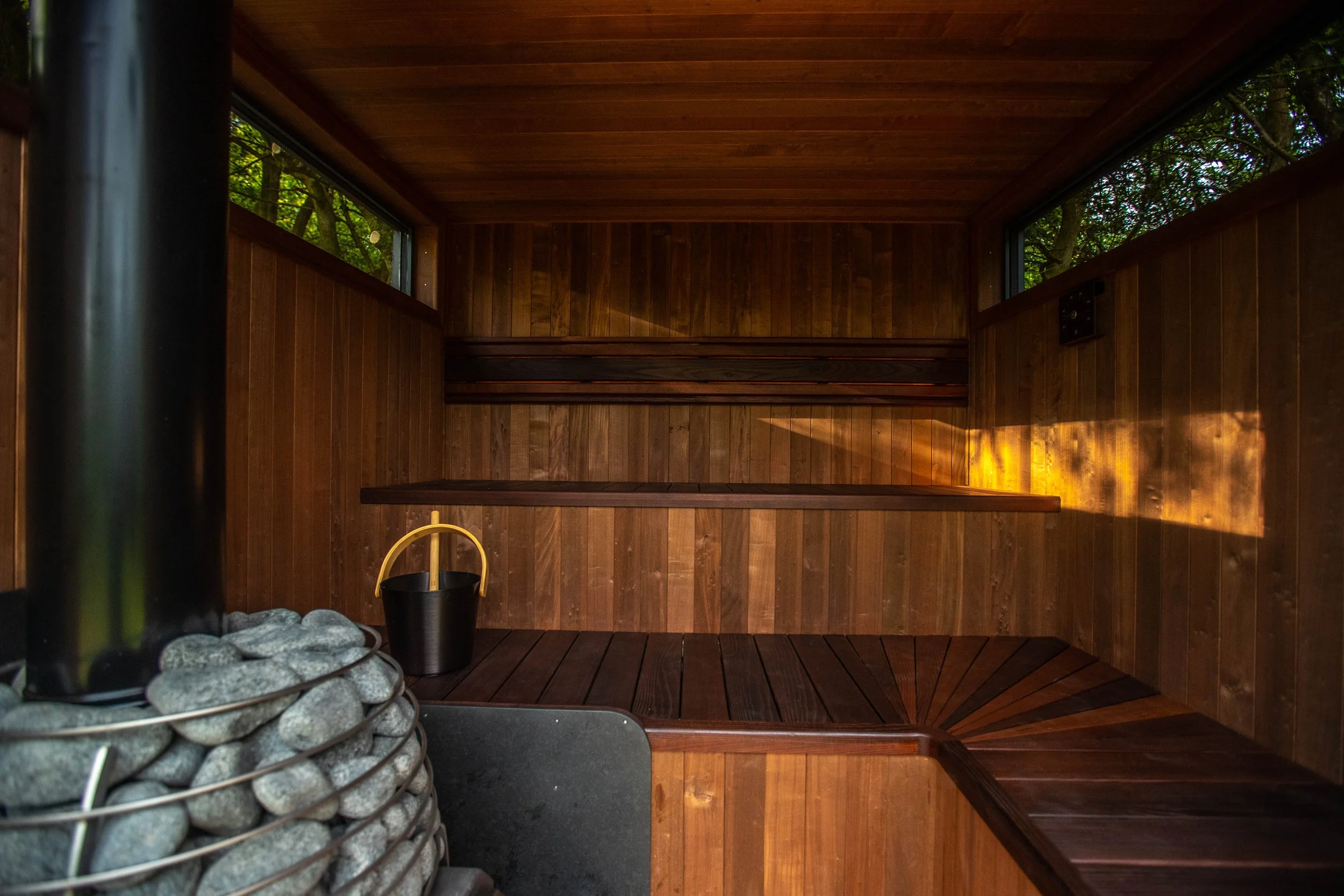 Interior of a wooden sauna with three tiers of benches, windows, and a heater with stones in the corner.