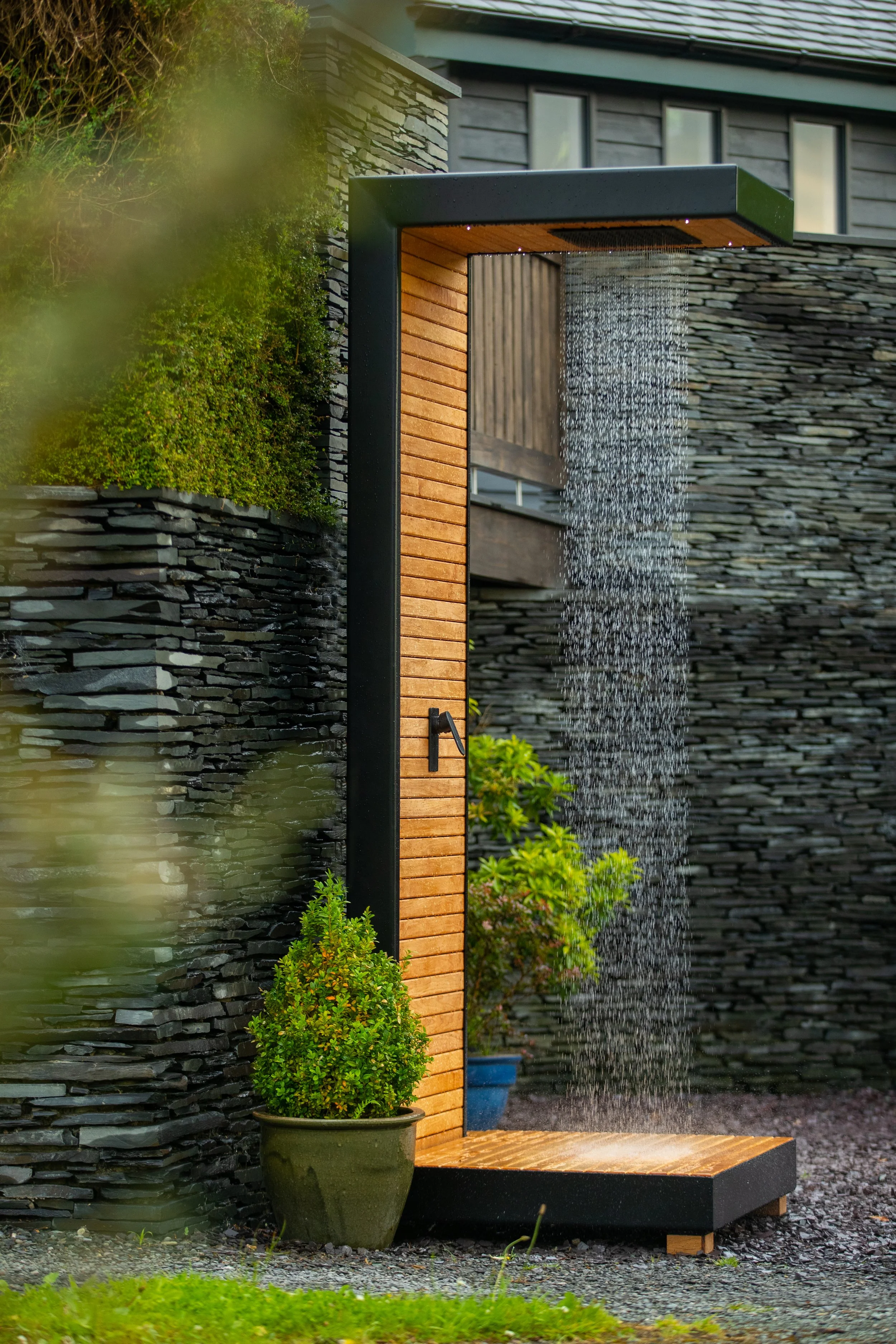 Modern outdoor shower with a rainforest shower-head, wooden panelling, black powder-coated metal frame, potted plants, and slate stone wall background.