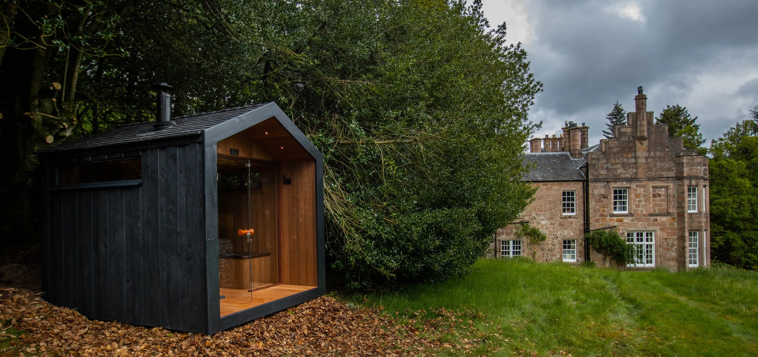 A modern black and wooden outdoor sauna with a glass door, situated on fallen leaves in a grassy area next to a large historic stone house with multiple chimneys and small windows, surrounded by trees under a cloudy sky.