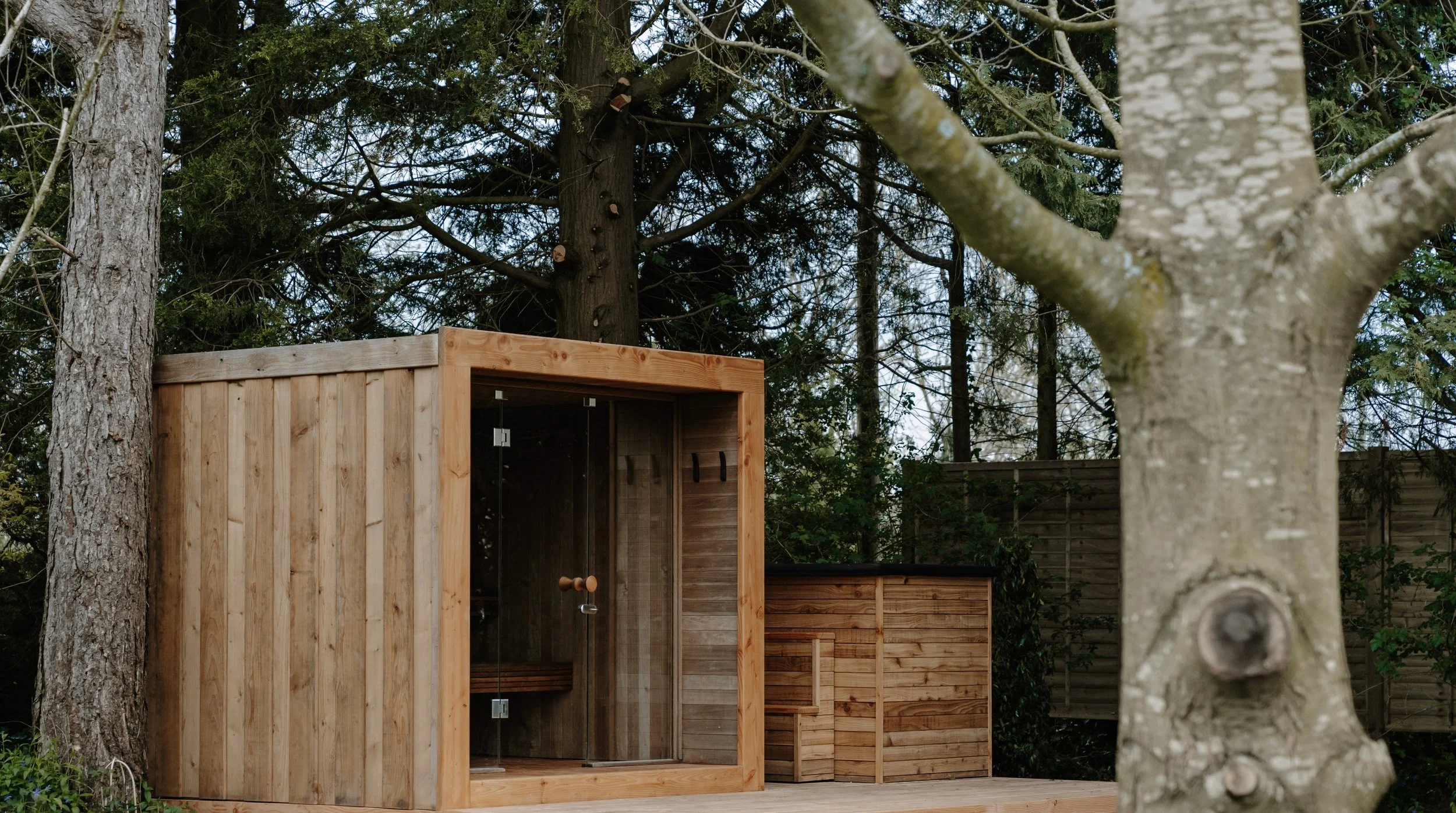 A wooden outdoor sauna with glass doors situated on decking with a wood-cladded plunge pool in the corner of a residential garden with trees in the background.