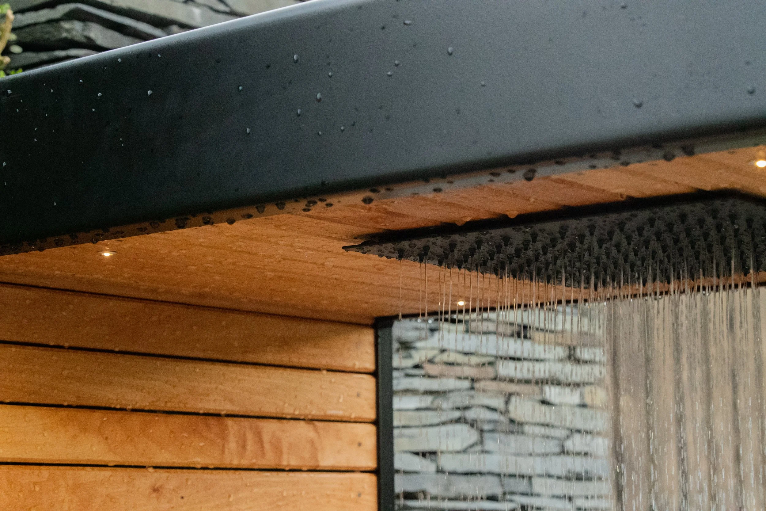 Water from a square rainforest shower-head falling on the edge of a modern outdoor shower with a black metal roof and natural wood-cladded wall.