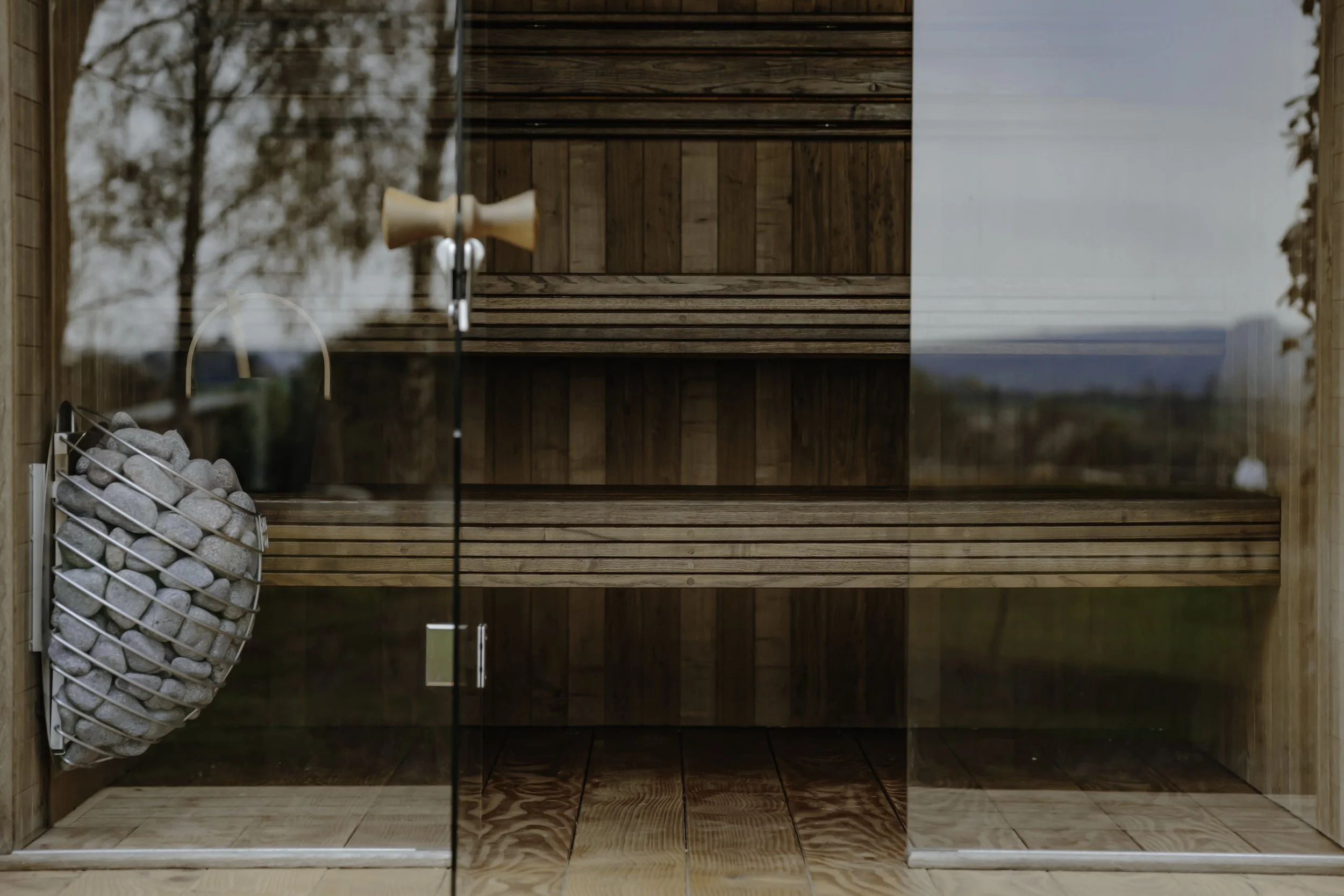 Inside a modern wooden sauna, with two tiers of bench seating and a heater filled with stones, viewed through glass panels, and outside scenery visible in the reflection.