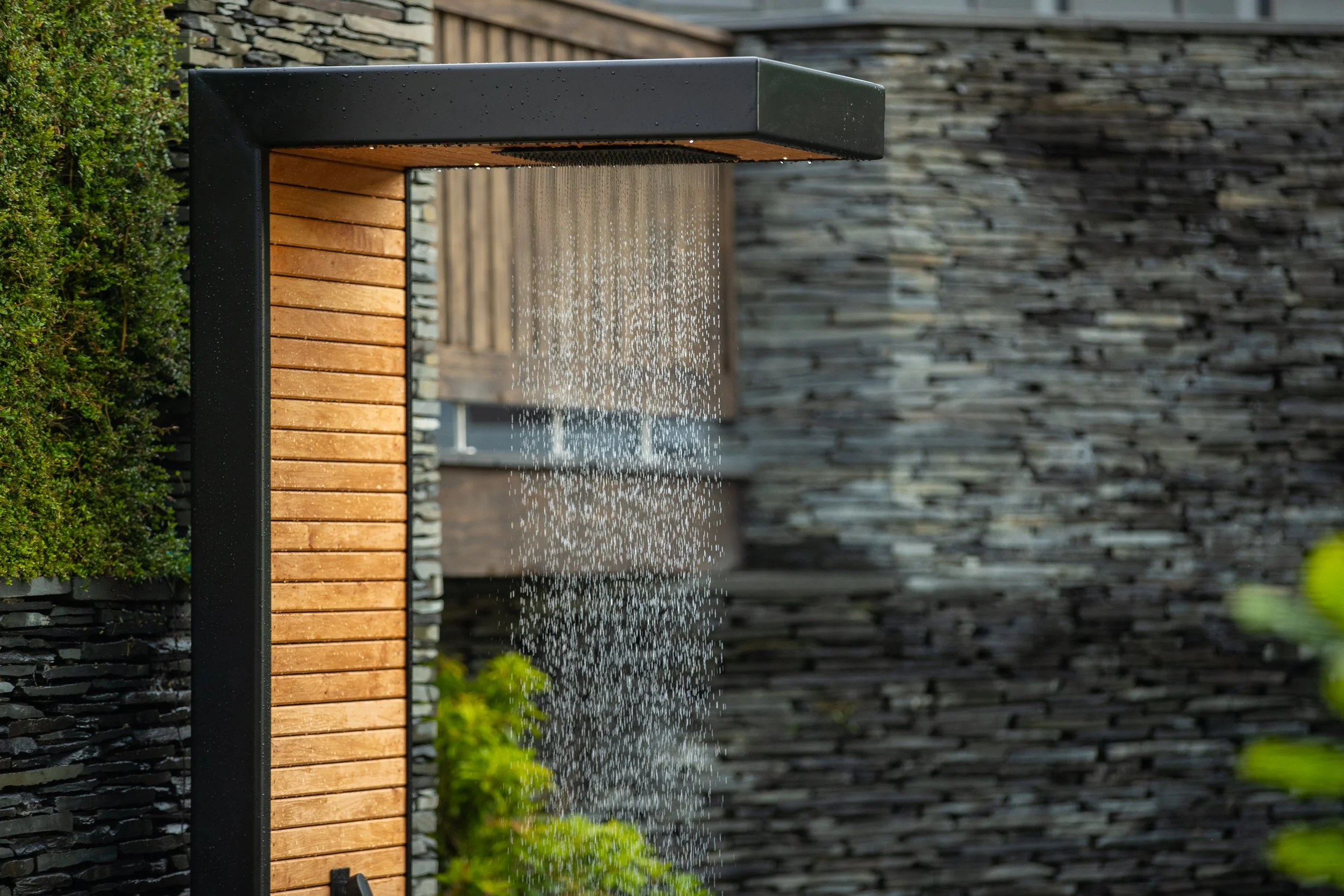 Outdoor shower of black metal and natural wooden cladding, with water cascading down, surrounded by a lush garden with greenery and a stone wall in the background.